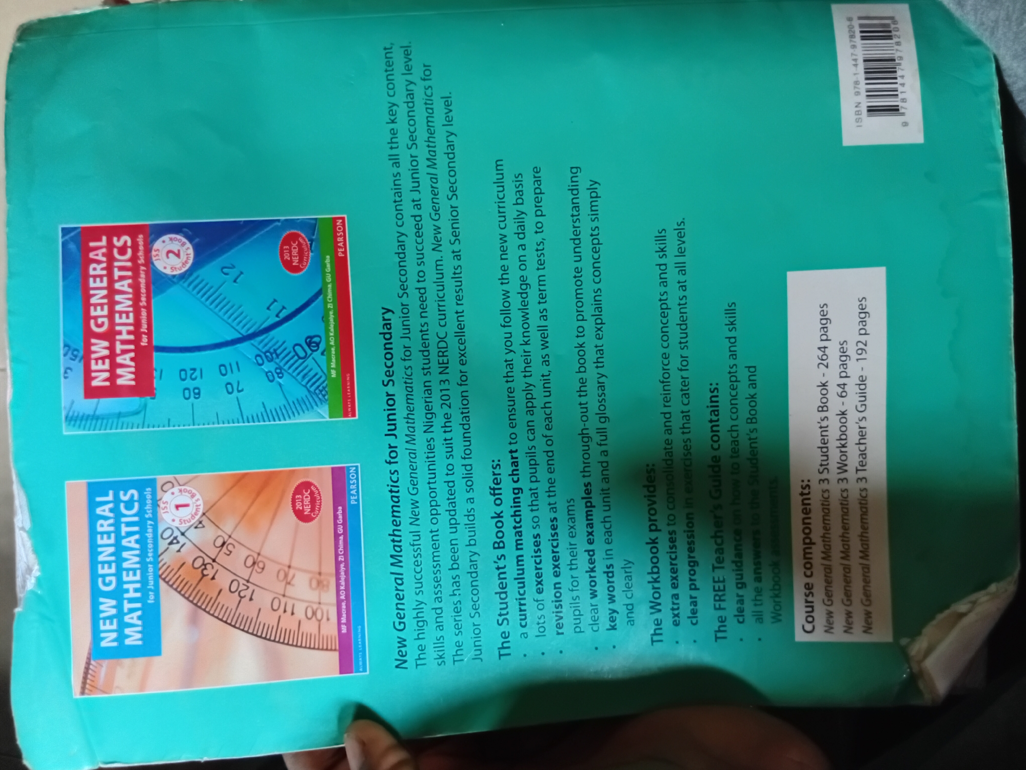 The image shows the back cover of a New General Mathematics for Junior Secondary Schools textbook, likely sold in Nkpor, Nigeria. The cover is predominantly teal green. Text on the cover describes the book's features, including "The Workbook provides," "The FREE Teacher's Guide contains," and "The Student's Book offers." There are also images of protractors and a "2013 NERDC Curriculum" sticker. In the bottom left corner, an ISBN barcode is visible with the number 978-1-4479-7820-6. The lighting suggests the photo was taken indoors with overhead lighting, creating some shadows and glare on the glossy surface of the book.