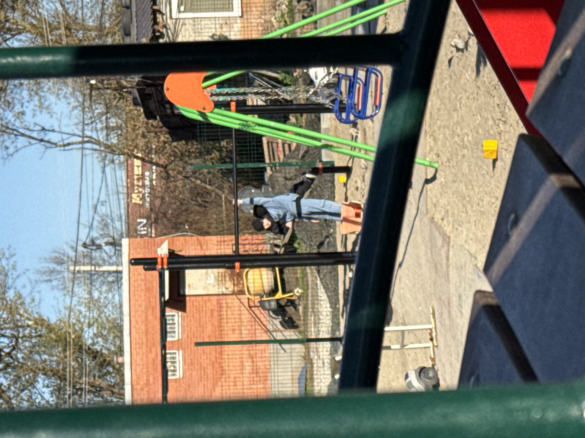 A young person wearing a martial arts uniform with a black belt is doing stretching exercises on the grounds of a playground. The playground contains typical play equipment: a metal swing set with two bucket seats and one metal swing, a yellow and orange slide, and a climbing structure comprised of green metal bars. A yellow building block toy lies on the ground near the slide. In the background are buildings, including a brick building with a white sign that reads "KY3HEU" in Cyrillic. The setting appears to be Rostov-on-Don, Russia.