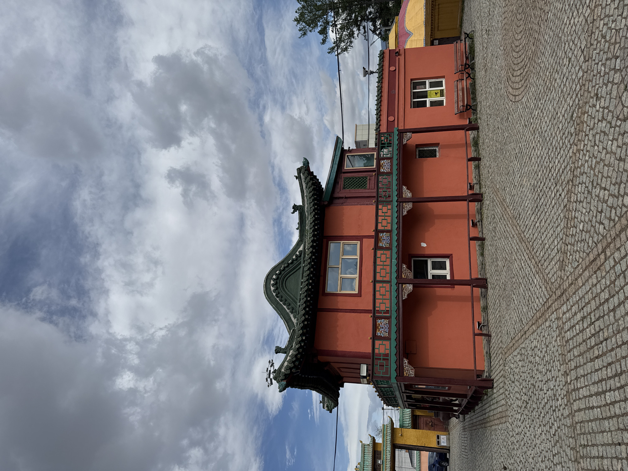 The image showcases a two-story, orange building with a distinct curved, green-tiled roof in Ulaanbaatar, Mongolia. The building's architecture features intricate wooden detailing and windows on both floors. A balcony extends from the second floor, adorned with decorative elements. The building stands on a cobblestone-paved area. The sky is partially cloudy. Electrical wires run across the scene.