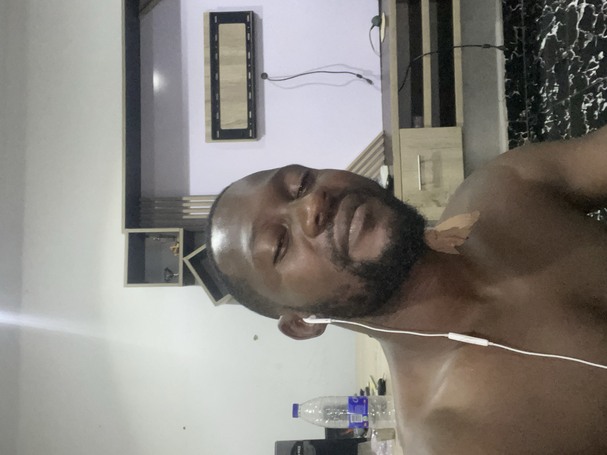 The image shows a close-up, low-angle, selfie-style shot of a Black man in his late 20s or early 30s. He has short, dark hair and a full beard. He is wearing white earbuds with a cord extending down his chest. A light brown bandage or adhesive strip is visible on his right collarbone. The man appears to be indoors, in a residential setting, likely in Abuja, Nigeria.
Behind him, a light-colored wall serves as the background. To the left, a portion of a black desk or shelf is visible, with a clear plastic water bottle and a small metallic object on it. Above the desk area, there are two light brown, geometric shelving units mounted on the wall. To the right of the shelves, a dark-framed, rectangular panel with a light-colored wood interior is mounted on the wall. Below this panel, a modern, light-colored entertainment center or console is positioned, featuring cabinets and open shelves. A black cord dangles from the wall panel and connects to a black power strip on the entertainment center. The floor in the foreground is covered with black and white patterned tiles. The lighting suggests an indoor, possibly evening, setting, with a strong light source casting a glare on the man's forehead and shoulder.