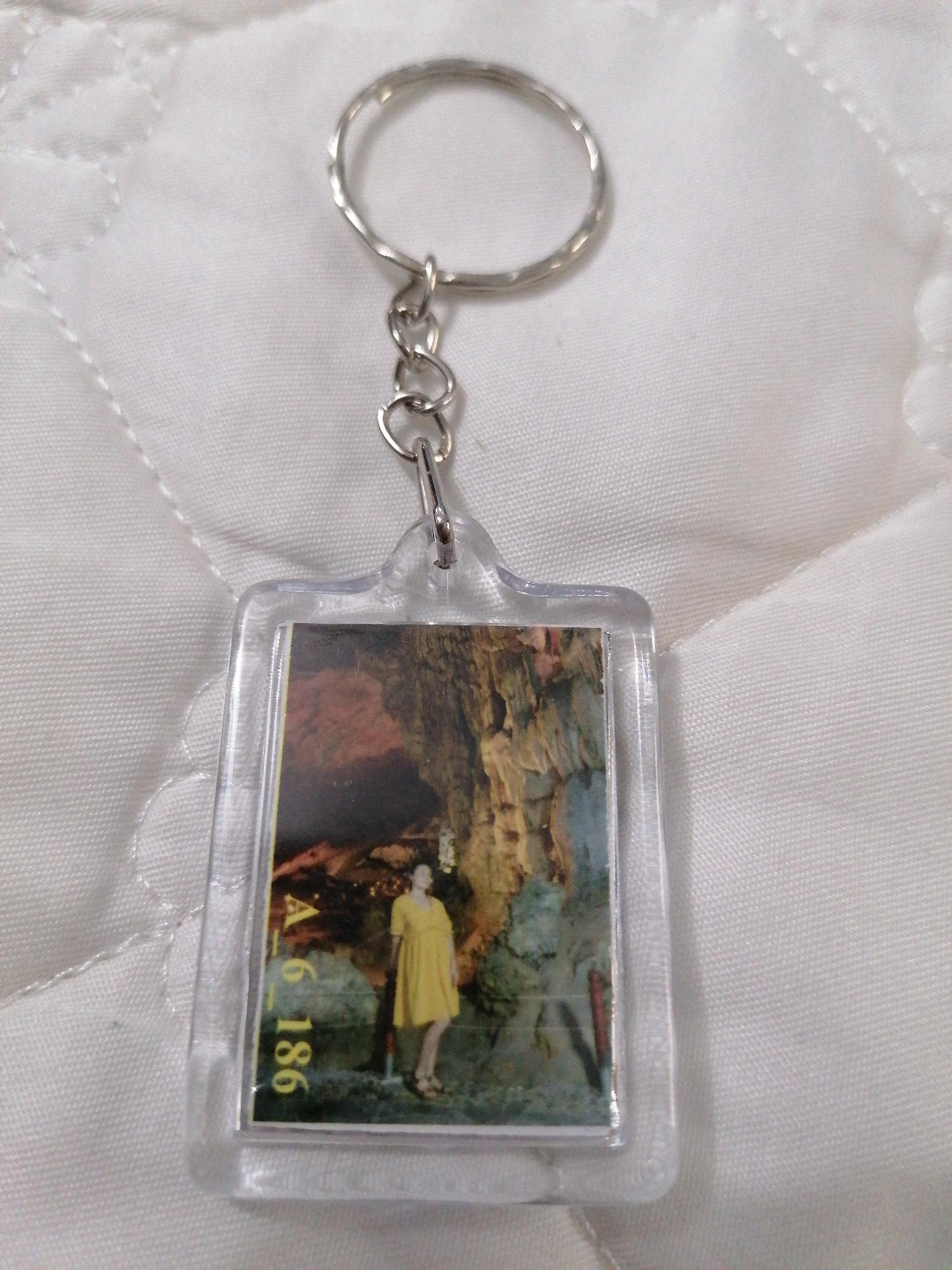 A silver-toned metal keychain with a clear rectangular plastic holder is centered on a white, quilted fabric surface. The keychain consists of a circular key ring attached to a chain that connects to the plastic holder.
Inside the plastic holder is a vertical photograph depicting a person, likely female, standing within a large cave. The person is viewed from behind, slightly turned to their right, and is wearing a bright yellow knee-length dress and sandals. Their gaze appears to be directed upwards or towards the impressive cave formations. The cave interior is dimly lit, with parts of the rock formations illuminated in warm earthy tones of brown, orange, and green, suggesting stalactites, stalagmites, or other geological features. The scale of the cave appears significant, dwarfing the individual.
Visible vertically on the lower left side of the photograph, within the plastic holder, is the text "A.6 186". The photograph likely serves as a souvenir, capturing a moment from a visit to a cave, consistent with the geological wonders found in areas such as Bai Chay Ward, Vietnam.
