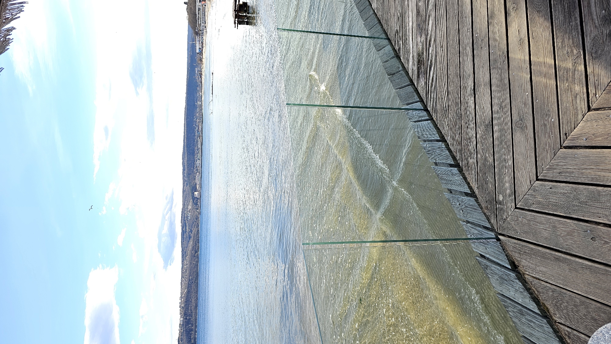 The image shows a coastal scene in Varna, Bulgaria. In the foreground is a wooden deck extending over the water. A glass railing separates the deck from the water. The water appears turbid, with visible wave patterns. In the distance, the Varna coastline with buildings and vegetation is visible. The sky is blue with scattered clouds.