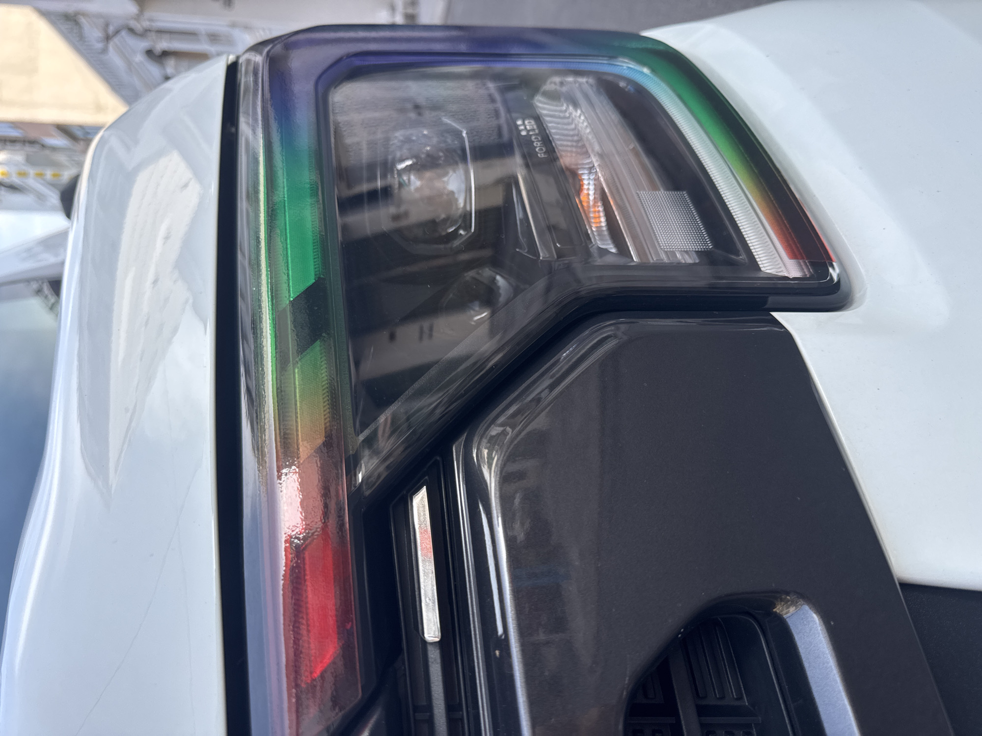 The image is a close-up, low-angle shot of the front right headlight and grille of a white Ford Raptor pickup truck, captured outdoors during the daytime. The headlight features a multi-colored LED strip, transitioning from red to green and then to blue, surrounding the main projector lens which is partially visible. The grille is black and features horizontal slats, with a subtle silver accent. The truck's white paintwork is visible on the hood and bumper. In the background, out of focus, are buildings and a street, suggesting an urban or suburban setting. The overall impression is a detail-oriented view of a modern vehicle's front design.