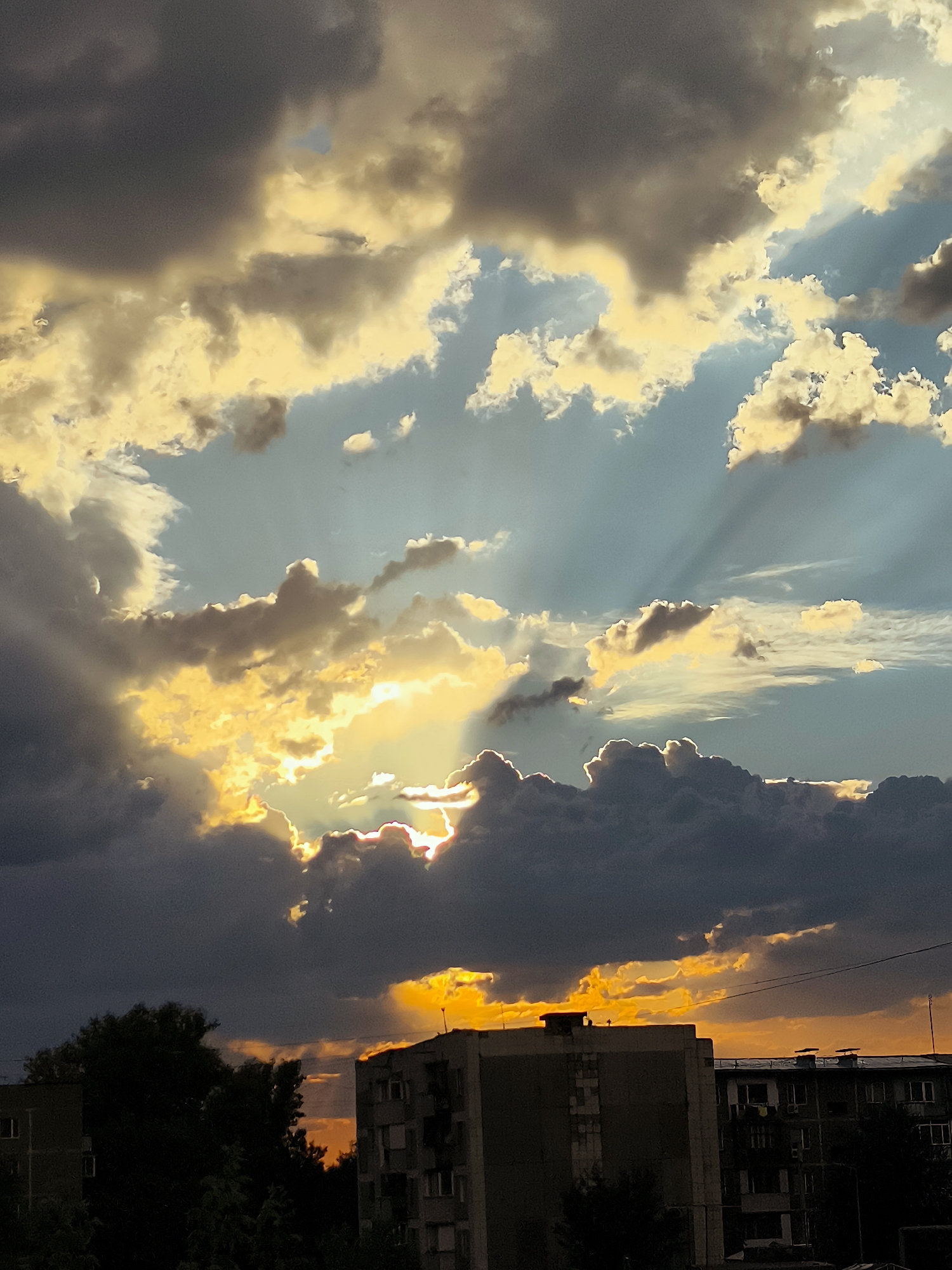 The image depicts a dramatic sunset over the city of Qarağandı, Kazakhstan. The sky is filled with a mix of dark, heavy clouds and bright, sunlit clouds that create a striking contrast. Sunbeams pierce through the clouds, illuminating portions of the sky with a golden hue. In the foreground, silhouetted buildings and trees suggest an urban environment. The architecture of the buildings appears to be multi-story residential blocks, typical of Soviet-era construction. The overall impression is one of a powerful natural phenomenon occurring over a populated area. The scene is captured from a slightly elevated perspective, looking towards the horizon.
