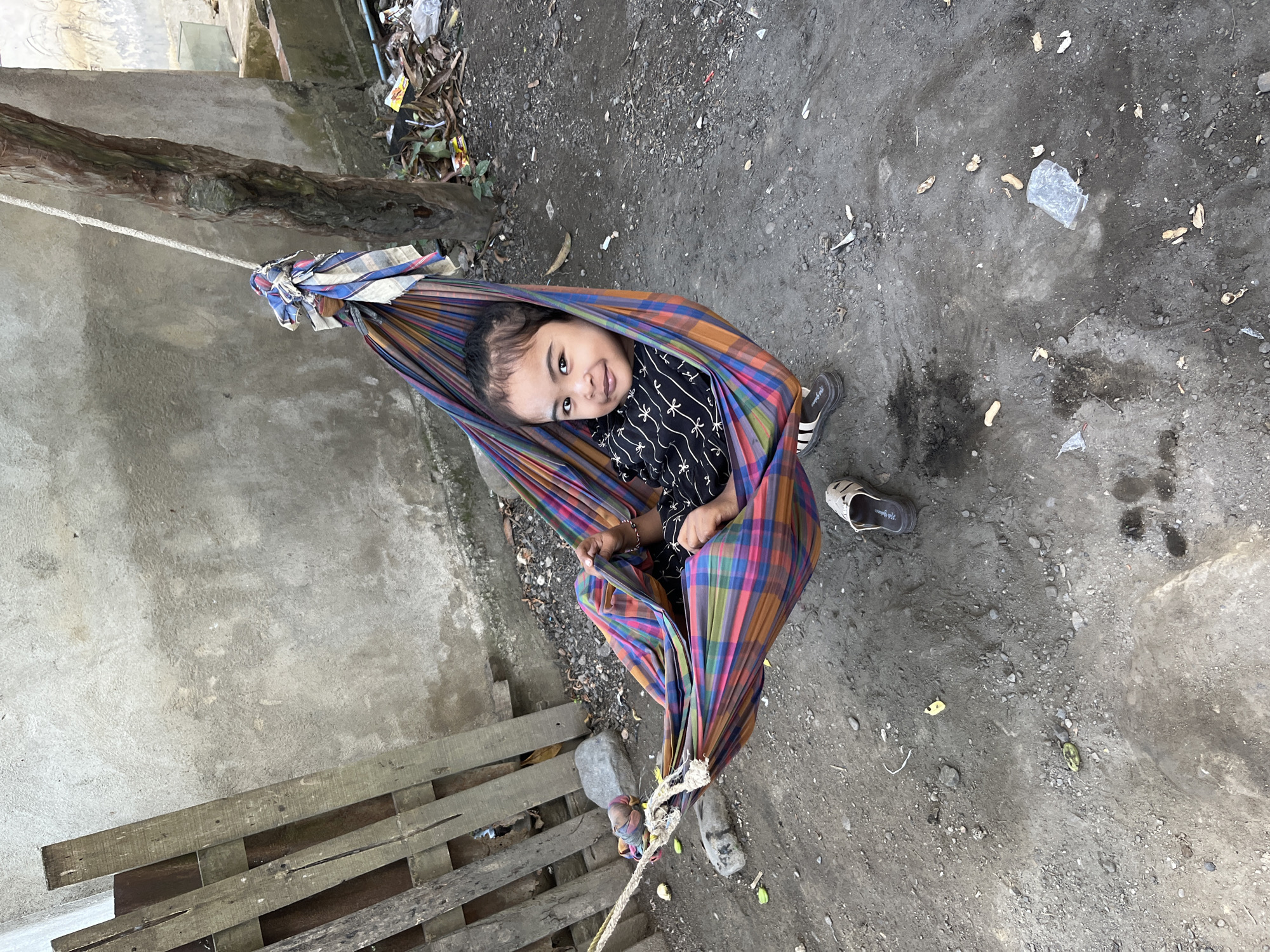 In Kruengmate, Indonesia, a young child is lying in a colorful striped hammock suspended between a tree and a wooden fence. The child is wearing a black shirt with a white pattern and appears to be smiling at the camera. The ground is dirt and gravel, with some debris scattered around. A pair of sandals lies on the ground near the hammock. A rough, concrete wall forms the backdrop, with a tree trunk visible on the right side of the frame.