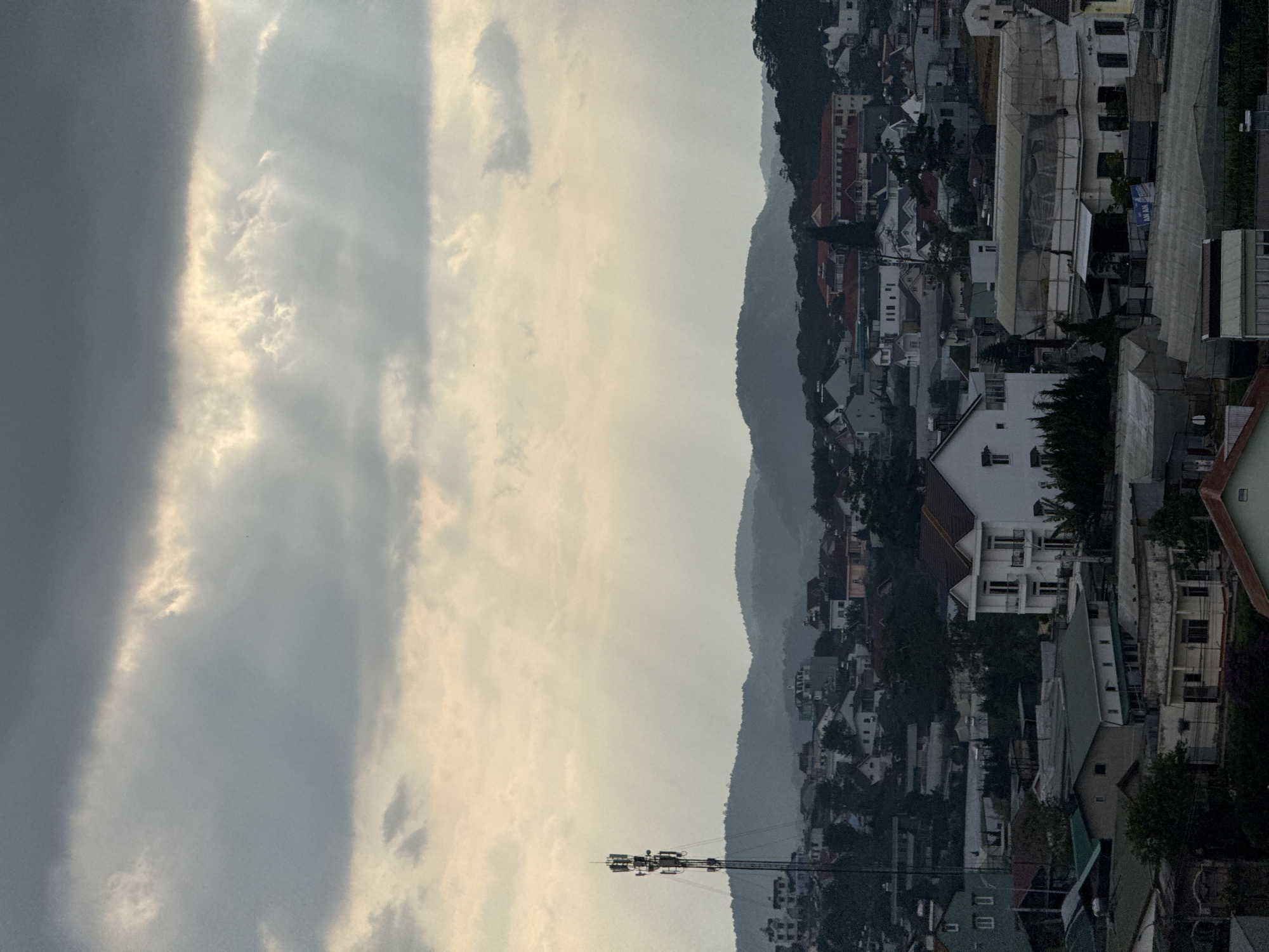 An urban landscape is captured against a backdrop of dramatic skies and distant mountains. The sky displays a range of cloud formations, from dense gray to brighter, sunlit patches. A communication tower stands prominently in the foreground. The cityscape below features densely packed buildings of varying heights and designs, with green spaces interspersed. The scene is likely in Lâm Viên - Đà Lạt, Vietnam.