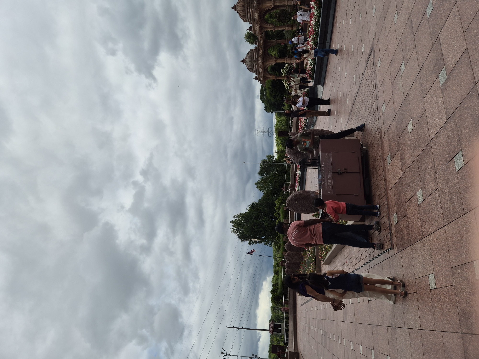 The image depicts the exterior of a religious site in Robbinsville Township, United States of America. The sky is overcast with heavy clouds. In the foreground, several people are gathered on a tiled plaza. A family, including two adults and two children, stand near a decorative metal box. To the right, another group of individuals are observing the surroundings, with some looking towards a building with ornate domes in the background. The environment features manicured flower beds and large stones. In the midground, an American flag is visible. Power lines and utility poles are also present in the background, suggesting an urban or suburban setting.