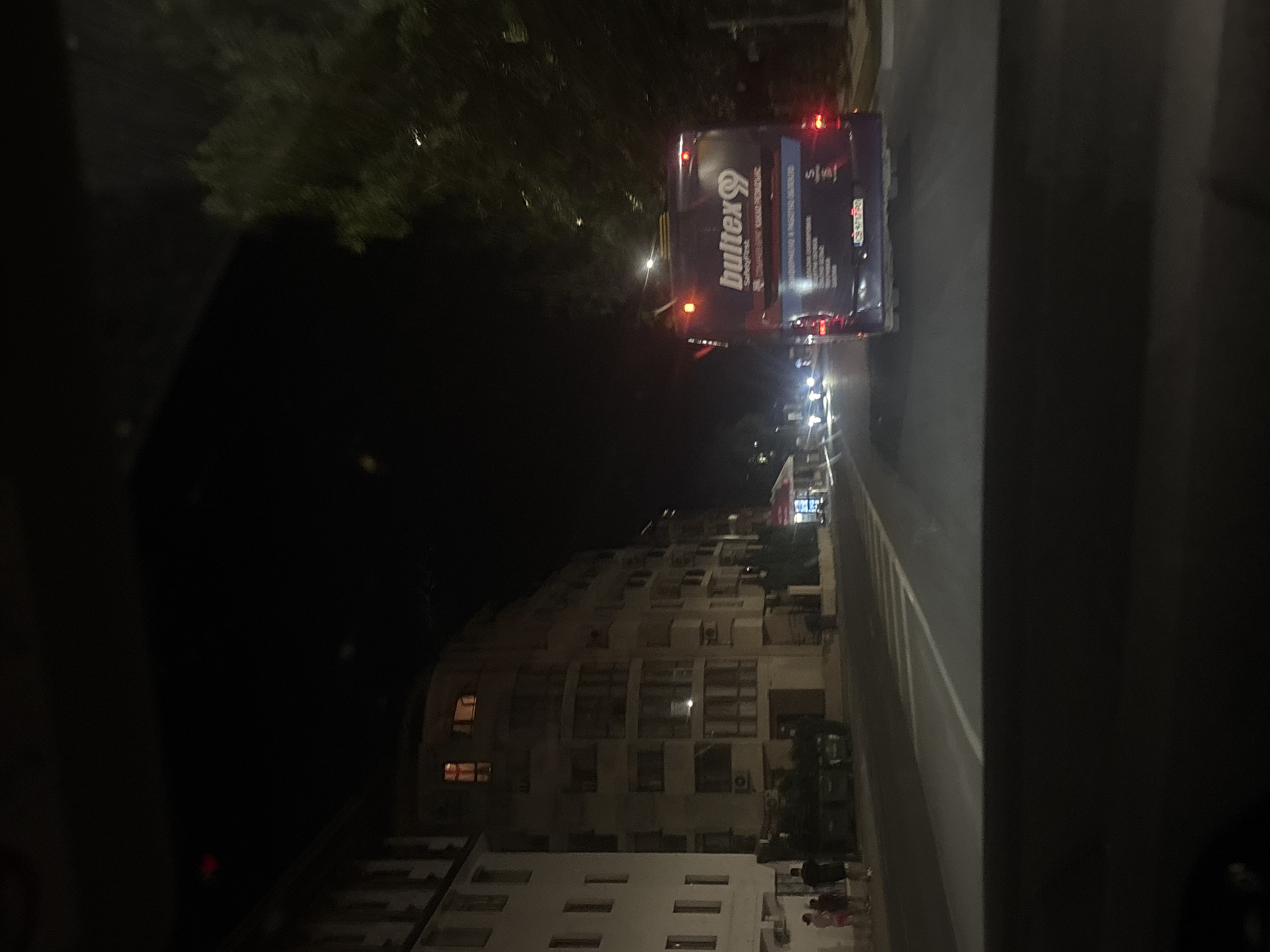The image captures a street scene at night in Nesebar, Bulgaria. In the foreground, the dark interior of a vehicle is visible, suggesting the photo was taken from a car. The road stretches ahead, marked with white lines. On the left side of the road, there are buildings with lit windows. Further down the street, a large blue bus with "bultex99" branding is driving away from the camera. The bus has its taillights illuminated. To the right of the bus, trees are visible. The overall atmosphere is dim, illuminated by streetlights and vehicle lights.