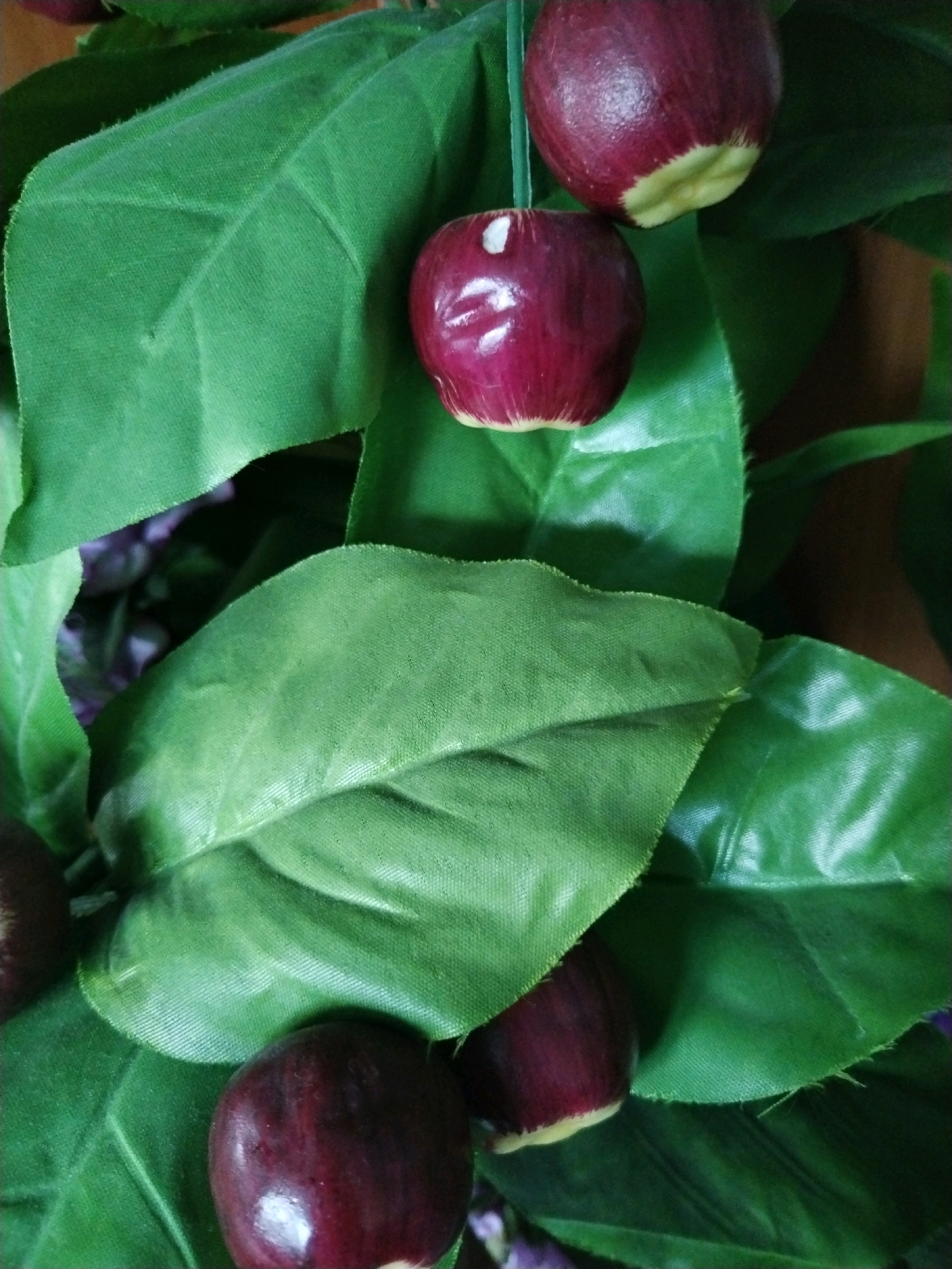 The image shows a close-up view of artificial cherries and leaves, arranged in a decorative manner. The cherries are a deep red or burgundy color with a yellowish-green stem end. They appear to be made of a smooth, glossy material. The leaves are a vibrant green, with visible veins and a slightly textured surface. Some of the leaves have a frayed edge. The overall impression is of a still life or a component of a larger artificial floral arrangement. There is no indication of any specific location in Baranavichy, Belarus, or any people present. The image is taken from a top-down perspective, with a shallow depth of field that blurs some of the background elements, creating a soft, naturalistic effect despite the artificial nature of the subjects.