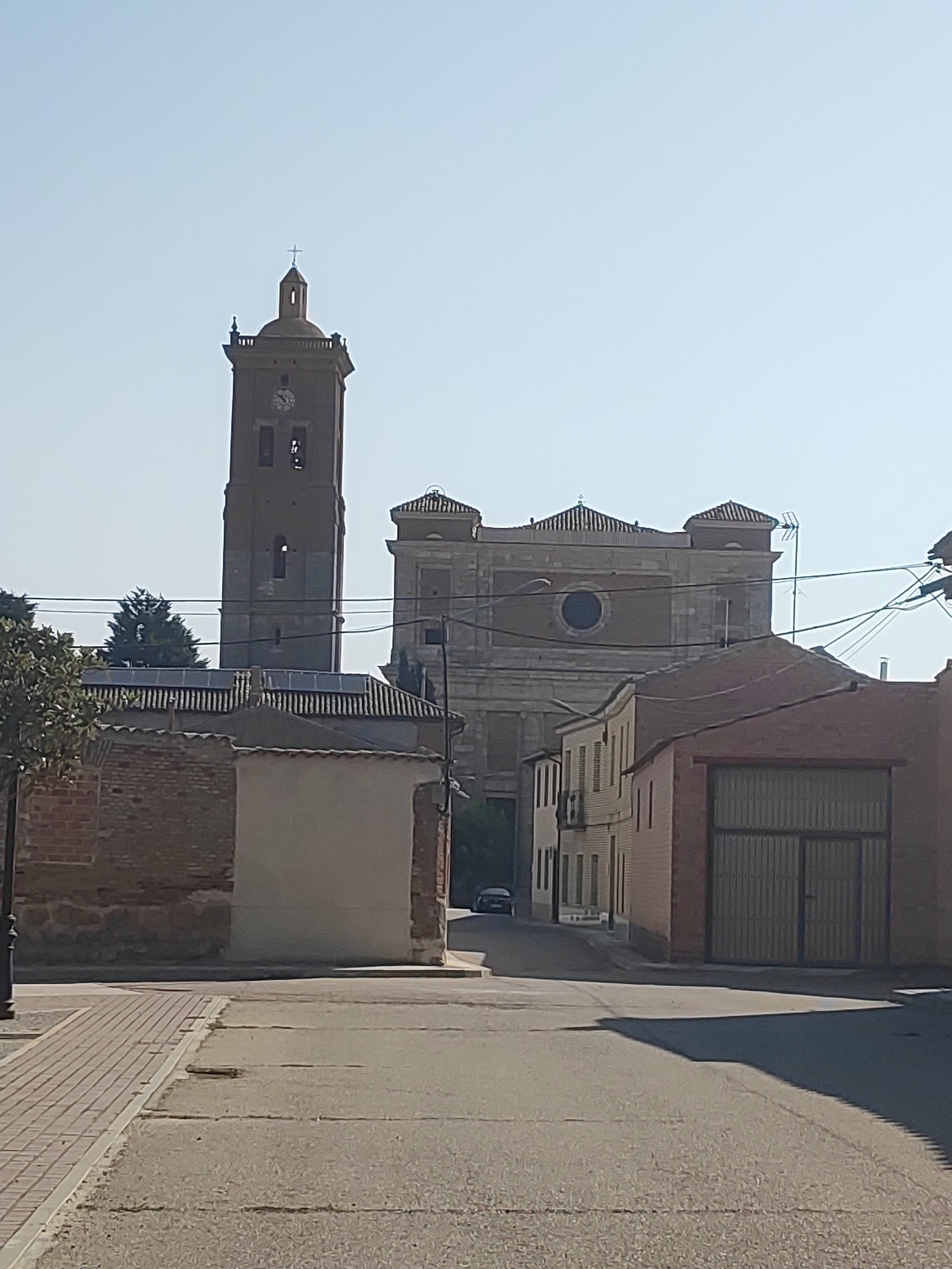 The image shows a street scene in Autillo de Campos, Spain. On the left, a brick wall with a lighter-colored section stands prominently. Beyond this, a tall, dark-colored church tower with a cross on top dominates the background, featuring a visible clock face. To the right of the tower, a large, light-colored building with arched windows and a circular opening is visible. In the foreground, a paved road leads towards the buildings, with a sidewalk on the far left made of light-colored brick pavers. A shadow of a tree or structure falls across the road from the right. The sky is clear and pale blue. A dark-colored car is parked on the street in the middle ground. Electrical wires are strung across the scene.