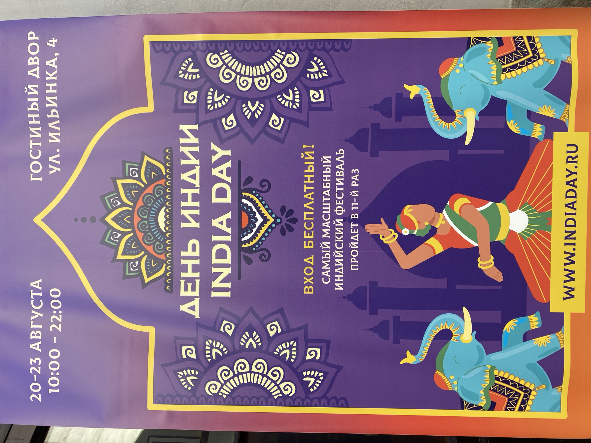 The image is a close-up of a banner advertising the "India Day" festival. The banner is predominantly purple with yellow and red accents. At the top, it displays event dates and times: "20-23 АВГУСТА" (August 20-23) and "10:00 - 22:00" (10:00 AM - 10:00 PM). Below this, it provides the venue: "ГОСТИНЫЙ ДВОР УЛ. ИЛЬИНКА, 4" (Gostiny Dvor, Ilyinka Street, 4), indicating a location in Moscow, Russia. The central text reads "ДЕНЬ ИНДИИ INDIA DAY" in large yellow letters. Further down, it states "ВХОД БЕСПЛАТНЫЙ!" (Admission is free!) and "САМЫЙ МАСШТАБНЫЙ ИНДИЙСКИЙ ФЕСТИВАЛЬ ПРОЙДЕТ В 11-й РАЗ" (The most massive Indian festival will take place for the 11th time). The bottom of the banner features a URL: "WWW.INDIADAY.RU". Visually, the banner is decorated with traditional Indian motifs, including mandalas on either side of the main text and stylized depictions of elephants and a dancer in the lower section. The dancer is shown in a vibrant red and green outfit, with hands raised in a traditional pose, and adorned with bracelets. Two blue elephants, decorated with colorful patterns, flank the dancer. The overall impression is festive and culturally specific, aimed at promoting an Indian cultural event.