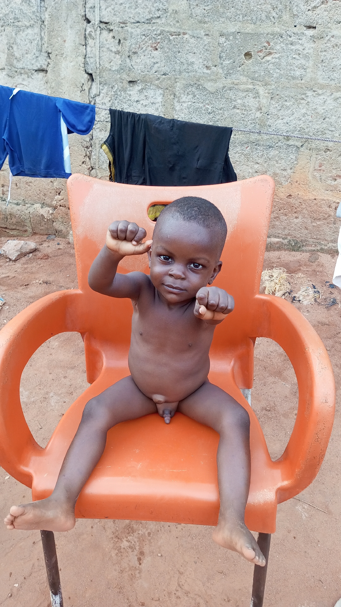 A dark-skinned, nude toddler sits on an orange plastic chair outdoors. The child's right arm is raised with a clenched fist, while the left arm points towards the viewer. In the background, a concrete wall is partially visible, with clothes hanging on a line above the chair. The ground appears to be reddish dirt. The image was taken in the city of Nkpor, Nigeria.