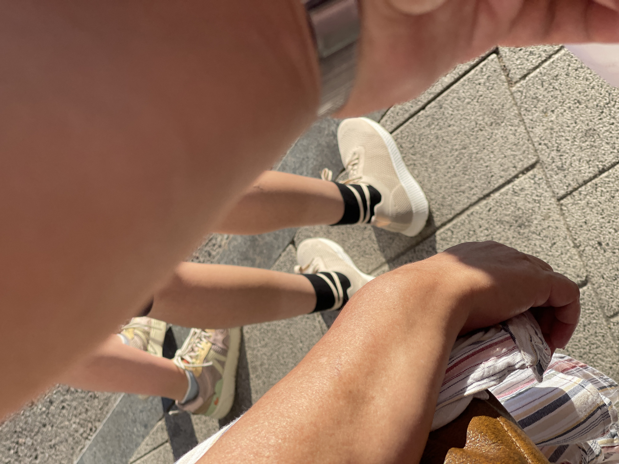The image captures a close-up view of legs and feet on a paved surface, likely in Zelenogradsk, Russia, given the context provided. Two individuals are partially visible. One person on the right wears light beige sneakers with black socks featuring two white stripes. Their leg is slender and appears to be in motion. Another person is seen on the left, whose arm, clad in a striped shirt, rests on a brown object. A second pair of sneakers, colored with pink, yellow, and green accents, is visible in the background, worn by someone whose legs are mostly obscured by the foreground. The ground is a light-colored pavement made of square tiles. The lighting suggests a sunny day.