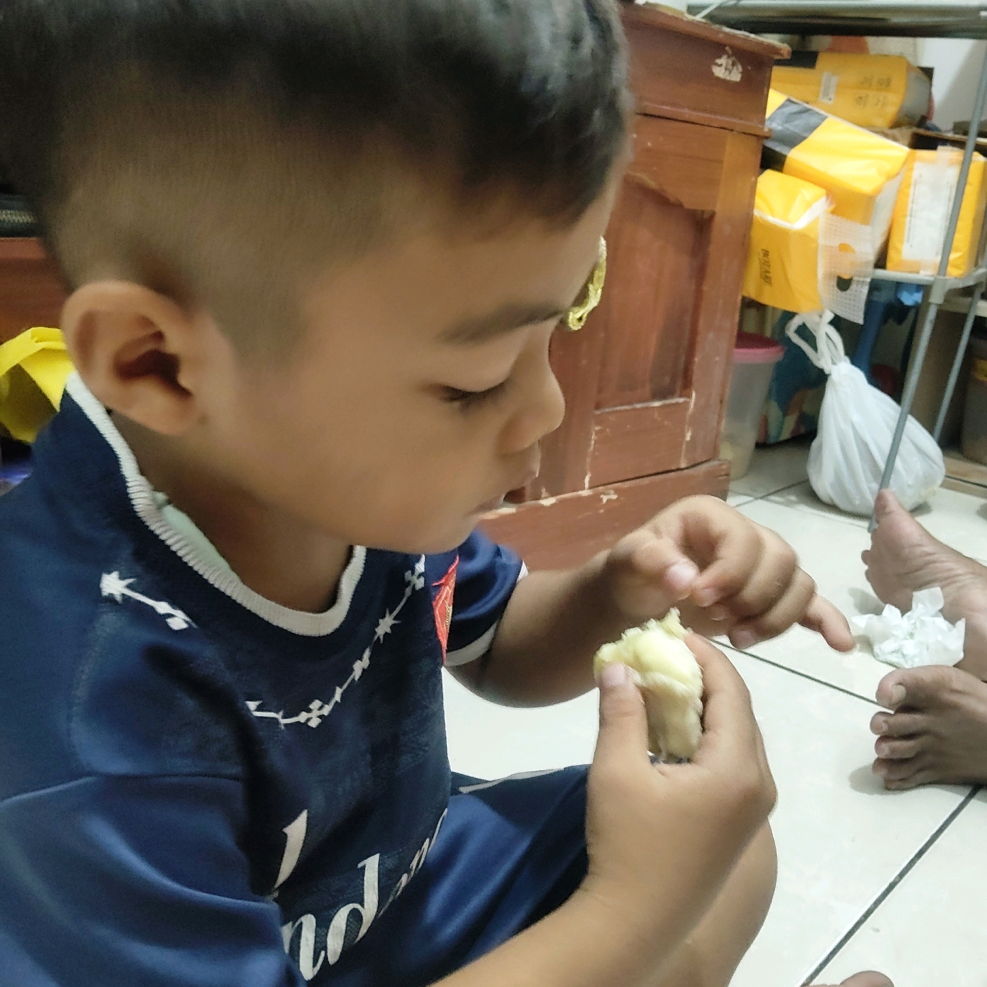 In Mekarmukti, Indonesia, a young boy in a dark blue shirt is eating a piece of food. He is seated on a tiled floor, with his hands holding the food. A portion of an adult's foot is visible to the right of the boy, with a crumpled piece of white paper near it. In the background, a wooden cabinet and shelves filled with yellow packaging are visible, along with a white plastic bag hanging from a rack.