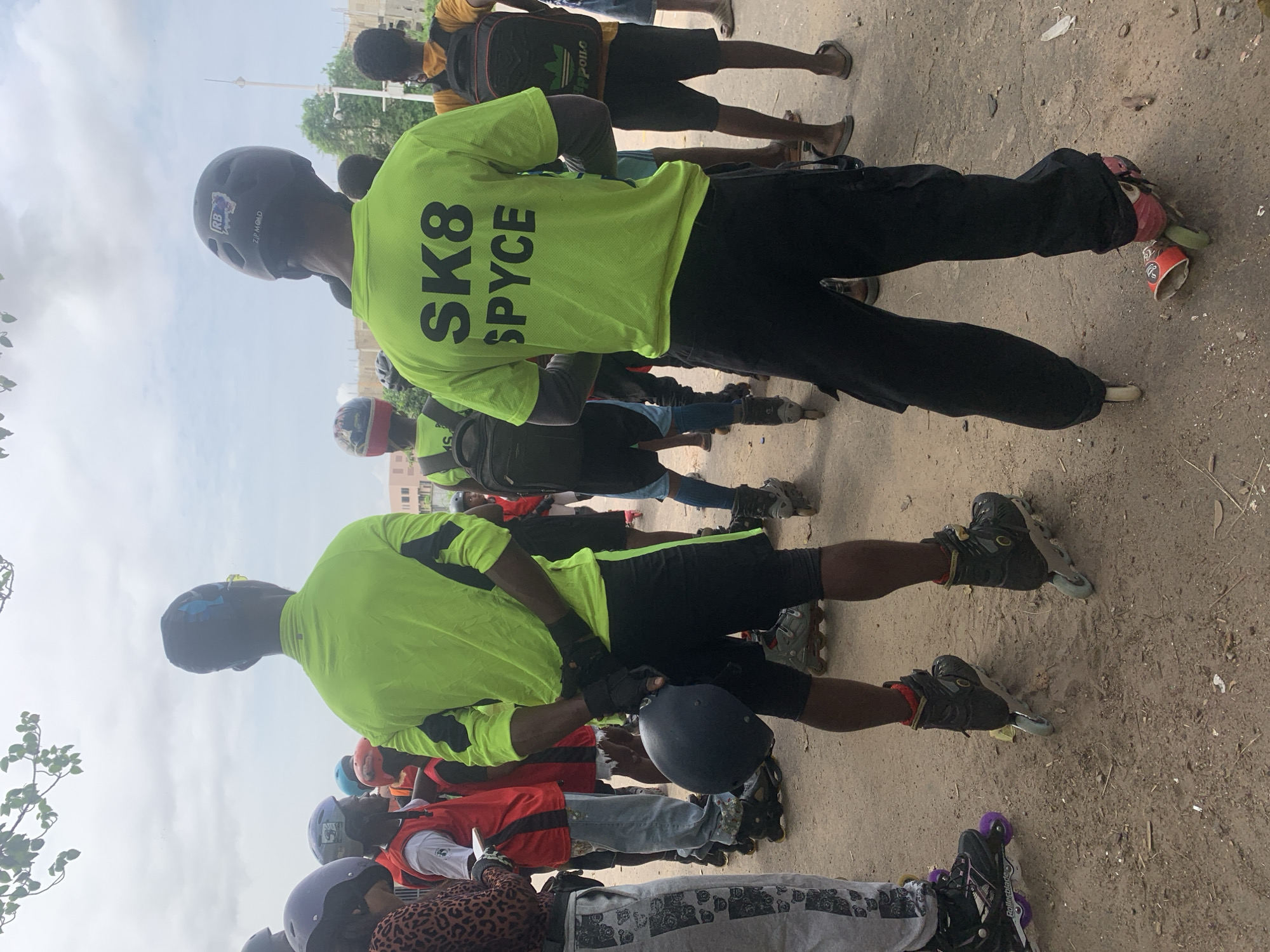 In Alagbede, Nigeria, a group of individuals, some wearing helmets and rollerblades, are gathered outdoors. The scene appears to be a dirt or gravel area, possibly a street or a public space, with a cloudy sky overhead. Several people are wearing bright, lime-green shirts with black lettering. One shirt prominently displays "SK8 SPYCE." Some individuals are holding helmets. The atmosphere suggests a gathering for a recreational activity involving rollerblading.