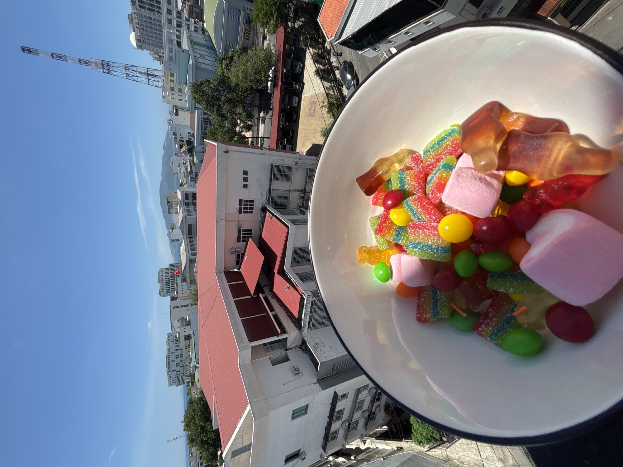 A bowl of colorful candies, including gummy bears, sour belts, and marshmallows, sits in the foreground, partially obscuring a view of Nha Trang, Vietnam. The candies are arranged in a white bowl with a dark rim. In the background, modern buildings with white facades and red-tiled roofs dominate the cityscape. A tall, lattice-like tower, likely a communication tower, stands prominently against a clear blue sky. The scene appears to be taken from a high vantage point, possibly a balcony or rooftop, offering a panoramic view of the city and the distant sea.