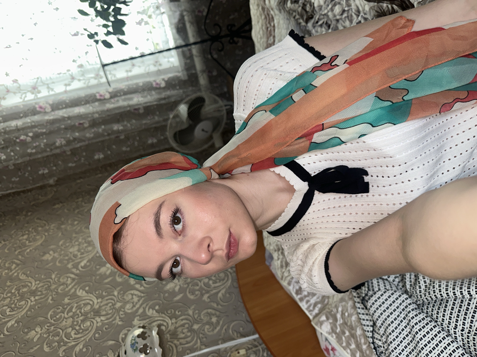 A woman wearing a headscarf and a white shirt with black trim is pictured indoors. The headscarf is patterned with colors including orange and teal. A fan is visible in the background near a window with a floral-patterned curtain. The woman is positioned on a patterned surface or bed, wearing a white shirt with black trim on the collar and sleeves. Part of her arm and the fabric of a garment with a black and white checkered pattern is also visible. The location, Коноваловка, Russia, is not discernible from the visual information.