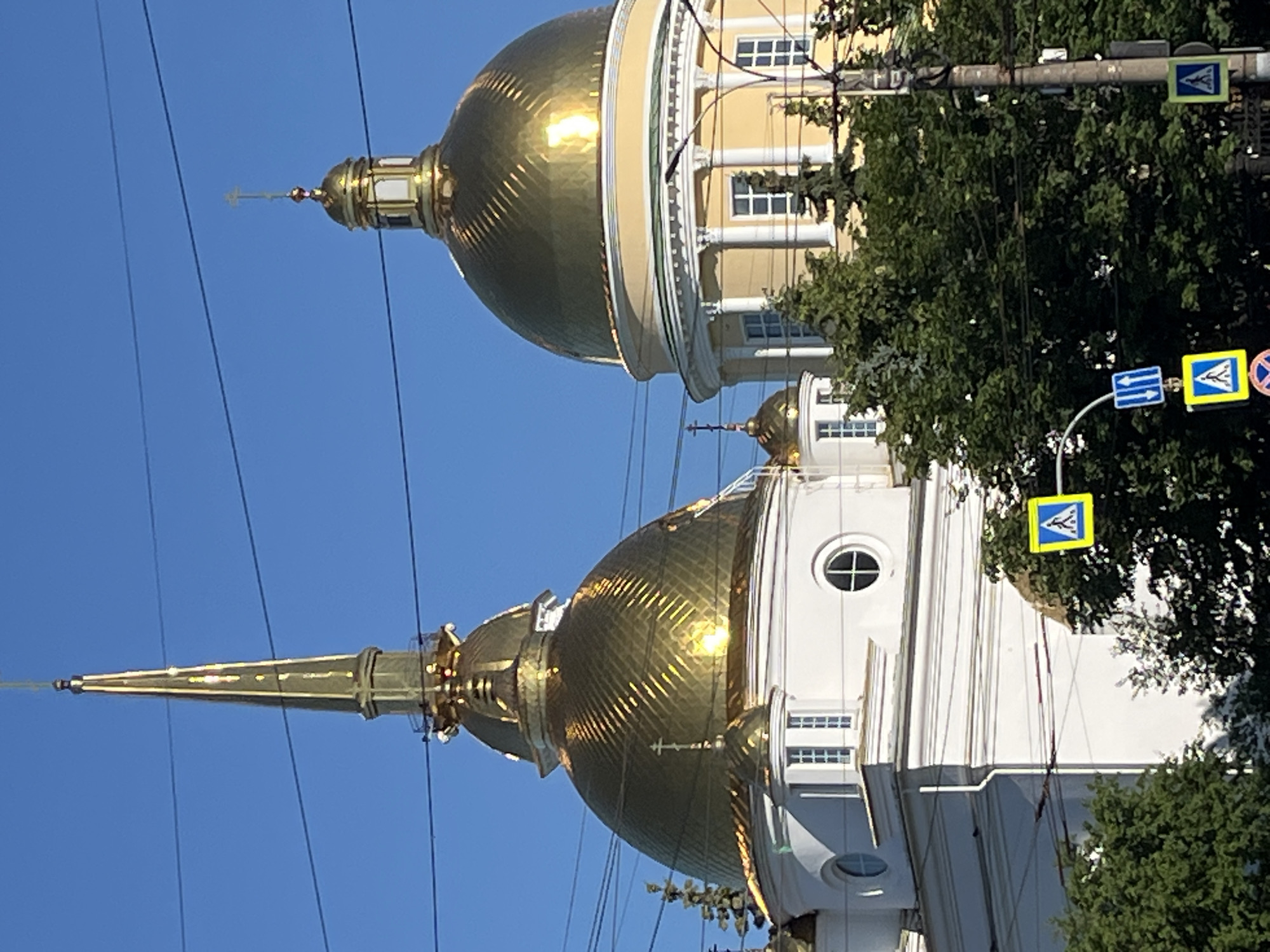 The image captures a close-up view of the golden domes and spires of the Saint Michael Cathedral in Izhevsk, Russia, under a clear blue sky. The architecture features classical elements with white and yellow facades. Several utility wires crisscross the frame, obscuring parts of the sky and the upper portions of the structures. In the foreground, lush green trees partially obscure the base of the cathedral, and various traffic signs are visible, including pedestrian crossing signals. The lighting suggests it is daytime, likely morning or afternoon, with sunlight glinting off the polished gold surfaces of the domes. There are no people or discernible actions taking place, presenting a static architectural scene.