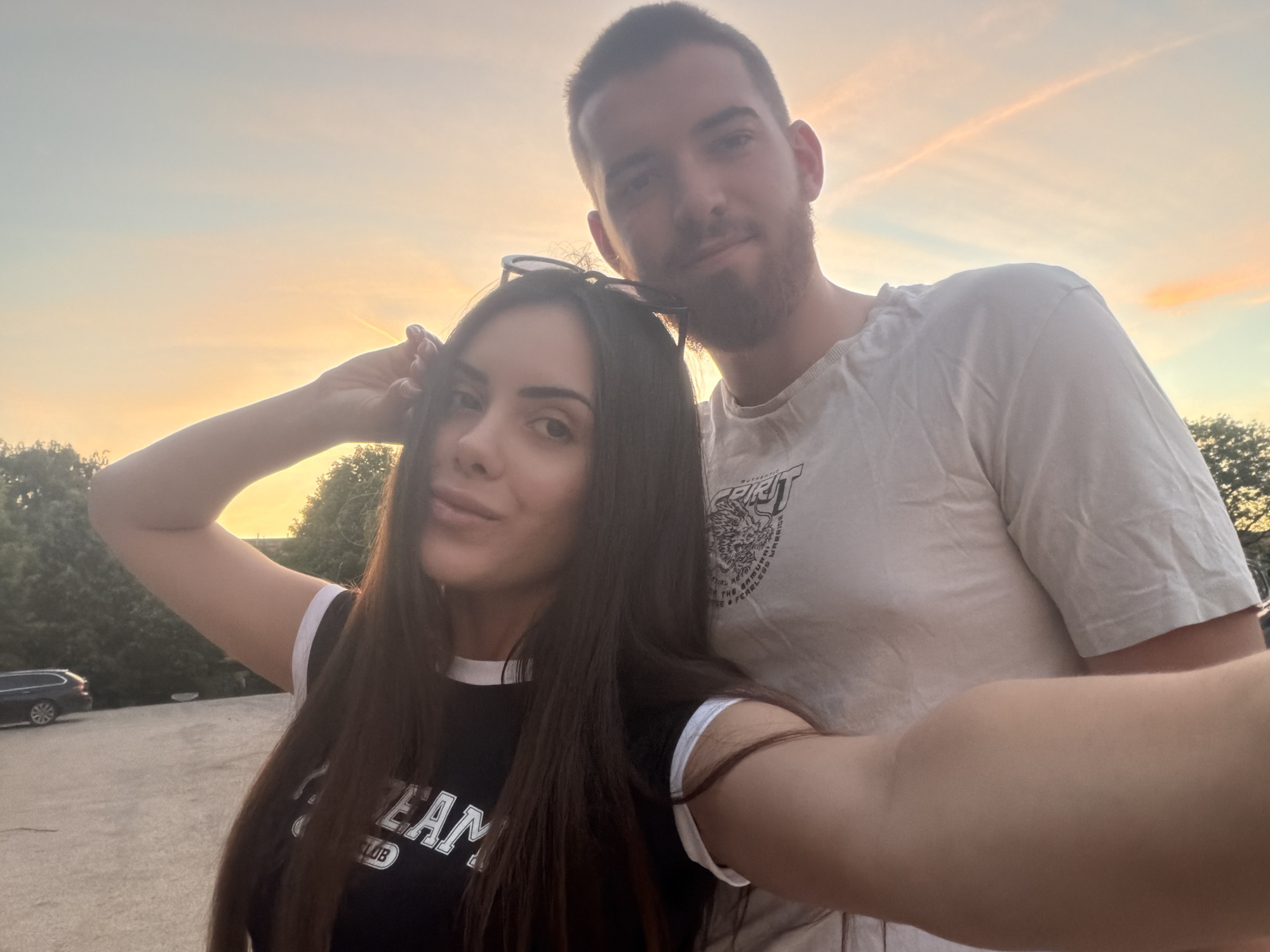 A young couple is posing for a selfie outdoors during golden hour. The woman, on the left, is wearing a black t-shirt with white trim and the text "TEAM CLUB" printed on it. She has long, dark hair and sunglasses on her head. She is raising her right arm. The man, on the right, has short brown hair and a beard, and he is wearing a white t-shirt. The setting appears to be a parking lot or outdoor area with trees in the background and a car parked on the left. The sky is a mix of orange and yellow hues, indicating either sunrise or sunset. While the location cannot be definitively identified, the city is provided as Sofia, Bulgaria.