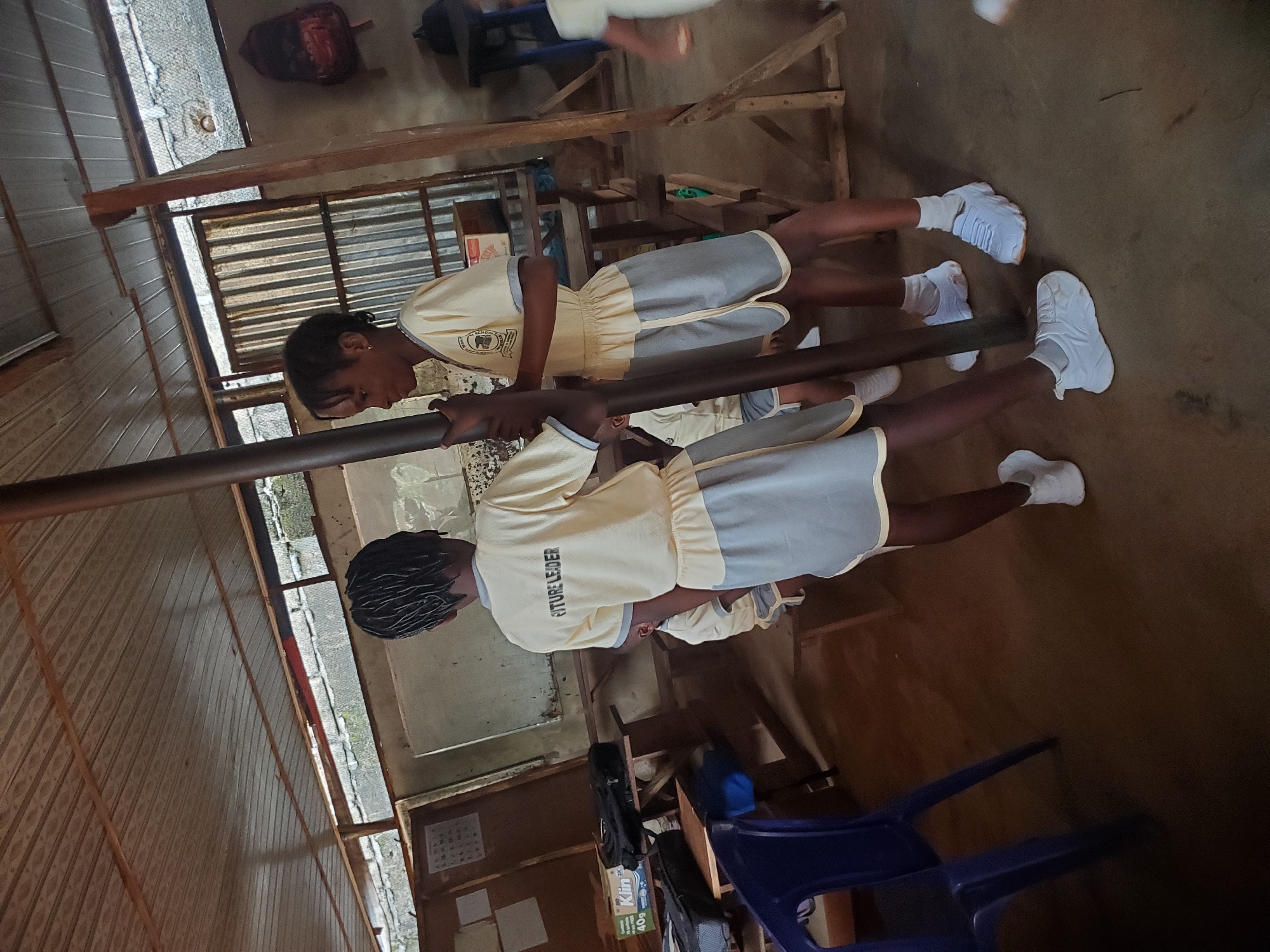 Two young individuals in school uniforms are gripping a metal bar. They are wearing white sneakers, grey skirts, and tan shirts with "FUTURE LEADER" printed on the back. The location is most likely Ukwu-Abwa, Nigeria. The scene takes place in what appears to be a simple classroom setting. The background contains wooden walls, corrugated metal roofing, desks, and a blue plastic chair.