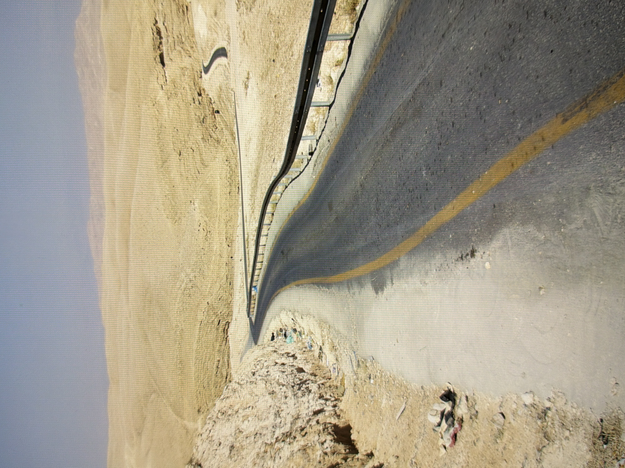 The image shows a winding asphalt road with a yellow dividing line, cutting through a desert-like landscape. On the left side of the road, there is a rocky, barren hillside with some debris scattered at its base. The road is bordered by a metal guardrail on the right, which curves with the road into the distance. The background consists of rolling, sandy hills under a pale blue sky. There are no people or vehicles visible in the immediate vicinity. This image does not depict any recognizable landmarks or features of the city of Kyiv, Ukraine.
