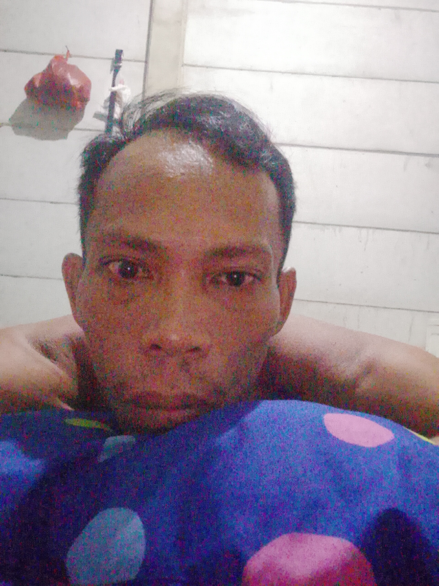 The image shows a person lying on a blue pillow with pink and white polka dots. The person's head is resting on the pillow, with their shoulders visible behind the head. The person has short, dark hair and a light complexion. Behind the person, there is a white wall with horizontal planks. In the upper left corner, there is a red object hanging from the wall. This image was taken in Bukit Ketok, Indonesia.