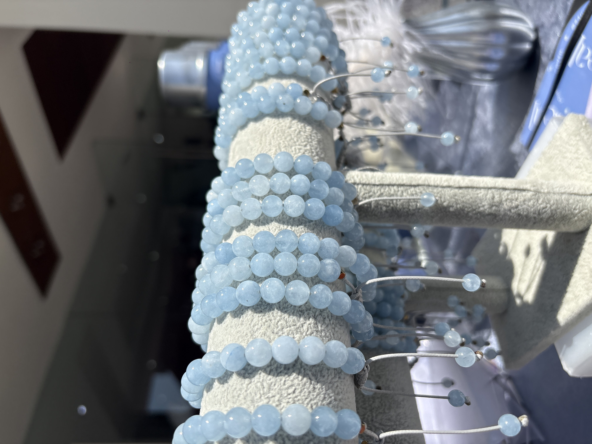 The image depicts multiple beaded bracelets made of light blue beads displayed on a gray, textured cylindrical stand. Each bracelet is threaded with a white string. Some bracelets have additional beads attached to the string. The scene is set outdoors, likely in daylight. The background includes hints of a white feather-like decoration, gray display elements, and blurred shapes indicating an indoor setting or display case. Based on the metadata, the location is San Andrés Cholula, Mexico.