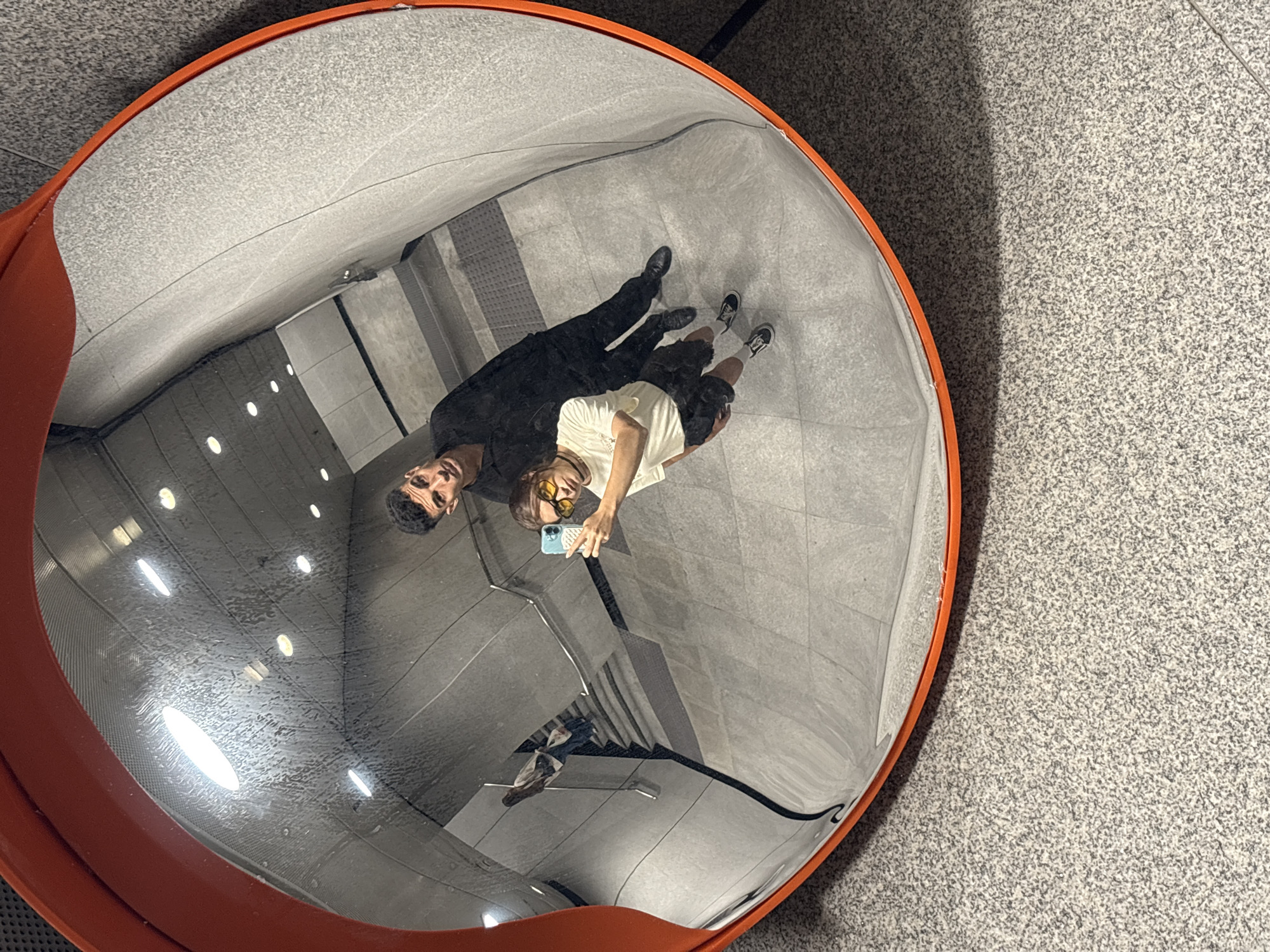 The image shows a convex mirror reflecting a man in a black suit, and another person wearing a light-colored shirt and shorts, holding a phone, likely taking a picture. The reflection includes a staircase, a wall, and ceiling lights. The area surrounding the mirror appears to be a gray tiled floor. The location is Hong Kong, China, based on the presence of a specific convex mirror design commonly found in that city.