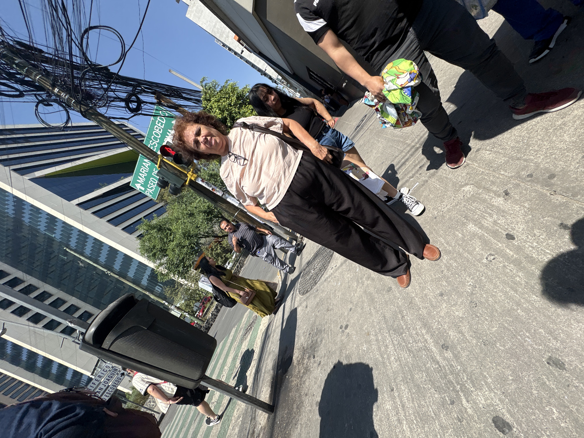 The image presents a bright, sunny urban street scene, likely in Mexico City, captured from a slightly tilted perspective. Several people are visible walking on a concrete sidewalk. In the foreground, an older woman with curly brown hair, wearing a light-colored t-shirt, black pants, and brown shoes, looks directly at the camera. Glasses hang around her neck.
Behind her, a younger woman with long dark hair, dressed in a black top and denim shorts, walks alongside another person in a black t-shirt and dark pants who is carrying a colorful, bundled item. Further down the street, other pedestrians include a man in a striped shirt and a woman in an olive-green dress.
A prominent utility pole, heavily tangled with numerous black electrical and communication cables, stands against a clear blue sky. Attached to this pole are green street signs and a traffic light displaying a red signal. In the background, a modern glass-faced building with a distinctive patterned facade is visible. A dark grey public trash bin is situated on the sidewalk.
The strong, distinct shadows cast by the people and objects indicate a clear, sunny day, likely midday or early afternoon. Visible text on the green street signs identifies major intersections: "MARIAN ESCOBEDO," "PASEO DE LA REFORMA" (partially visible), "PERIFERICO," and "SAN JOAQUIN ESCOBEDO." A section of the sidewalk features green and white painted stripes, characteristic of a crosswalk.