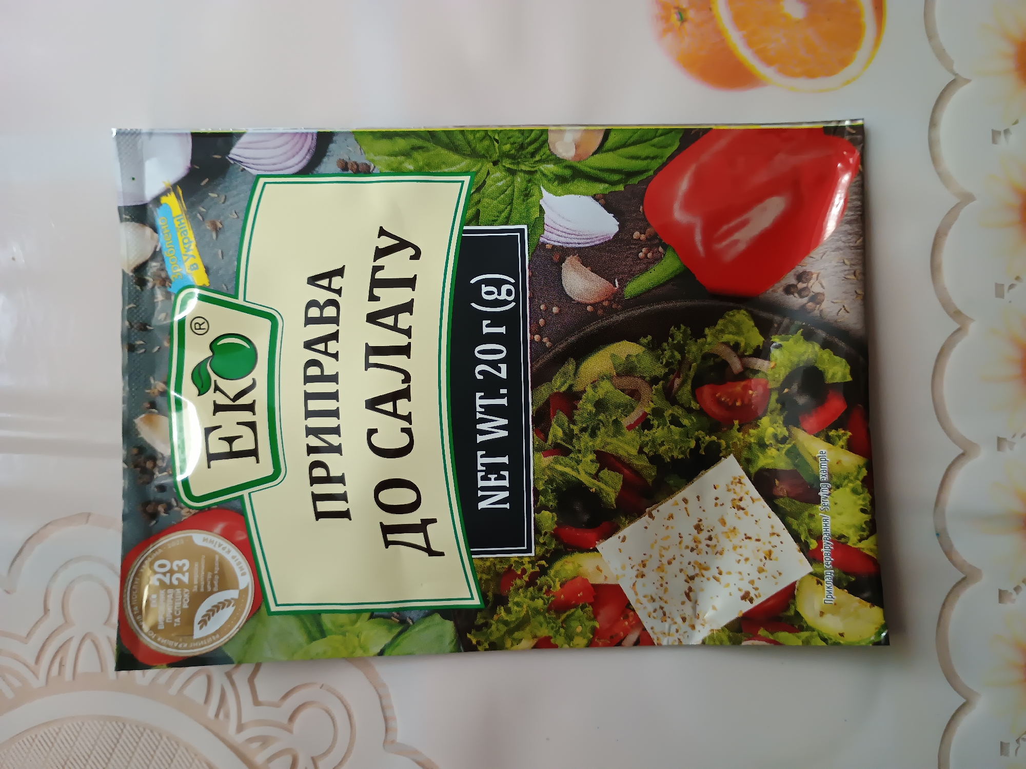 The image shows a package of seasoning for salad. The package features a picture of a salad with ingredients like lettuce, tomatoes, and cheese, as well as a red bell pepper. The text on the package is in Cyrillic and appears to be Ukrainian. There is a logo with an apple inside a circle. The text also includes "NET WT. 20g". There are also some oranges in the background. The image is taken from a top-down perspective.
