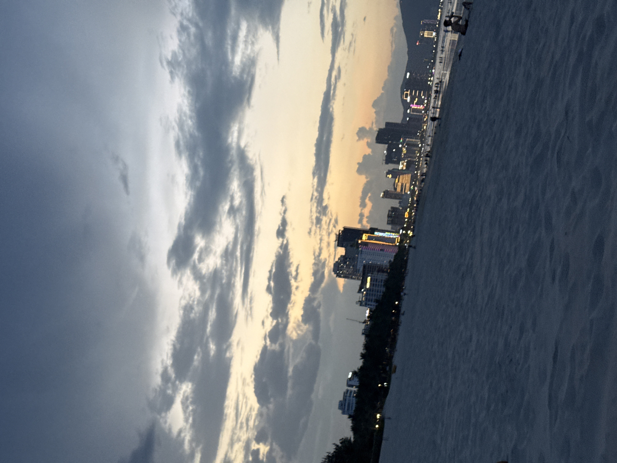 This image, when correctly oriented, presents a panoramic view of a coastal city at dusk or sunset. The sky, occupying the upper portion of the frame, displays a striking contrast: heavy, dark grey clouds dominate the left, while a radiant band of soft orange and yellow light, indicative of the setting sun, illuminates the right side. Lighter, scattered grey clouds traverse the mid-sky.
Below the expansive sky, the modern cityscape stretches horizontally, showcasing numerous high-rise buildings. Many of these structures are brightly lit, their windows and facades glowing with artificial lights, suggesting an active urban environment as evening descends. Faintly visible in the distant background, mountains rise behind the city's structures.
In the foreground, to the left of the city, a broad expanse of textured, light-colored sand forms a beach. Small, indistinct figures of people are visible along the shoreline, suggesting leisurely activity like evening strolls. The blend of natural light from the sunset and the vibrant artificial lights of the city creates a dynamic and atmospheric scene.
The setting aligns with Đà Nẵng, Vietnam, a city known for its beautiful beaches, modern architecture, and scenic mountain backdrops. There is no visible text in the image.