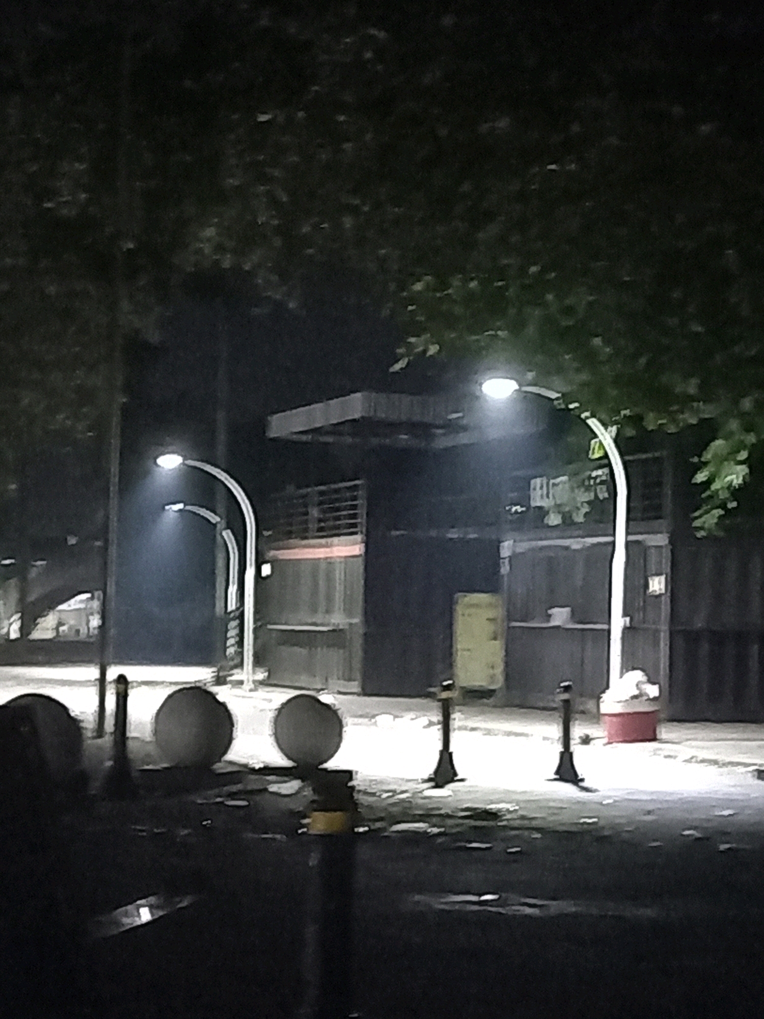 This is a nighttime, low-light, medium-distance, full-frame, static shot of an outdoor urban scene in Kandangjati Kulon, Indonesia. The image captures a street or pathway illuminated by streetlights.
In the foreground, there are several dark, circular objects, possibly bollards or decorative elements. A single, black pole is visible between two of these objects. Further back, a row of streetlights with curved poles illuminates the scene. The closest streetlight is on the right, with its light casting a glow upwards and outwards. Two other streetlights are positioned to the left, with the one nearest to the viewer showing a faint light.
Behind the streetlights, there are large, dark structures that appear to be buildings or shipping containers, some with visible doors or openings. One structure on the right has a red and white cylindrical object at its base. Yellow rectangular objects are visible against some of the structures. The ground is paved and wet in some areas, reflecting the light. Trees with dense foliage are visible in the upper portion of the image, obscuring the sky. The overall impression is one of an industrial or commercial area at night. There are no people visible in the image.