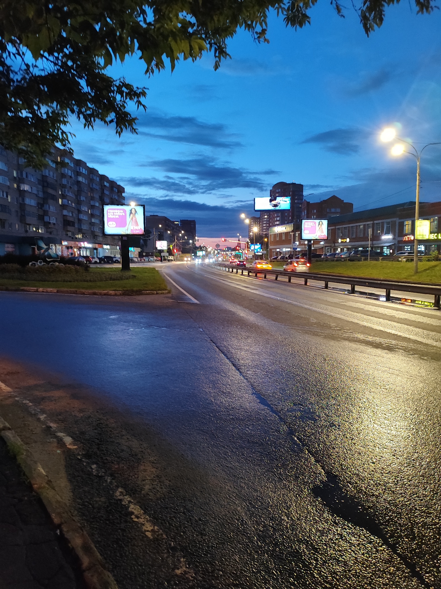 The image captures a city street at dusk or early night, with a clear blue sky showing some clouds. The street is wet, reflecting the light from streetlamps and vehicle headlights. On the left side of the road, there is a multi-story apartment building with windows lit from within. In front of the building, there are some parked cars and a grassy area with a small hedge. Several billboards are visible along the roadside, displaying advertisements. One billboard shows a woman in a pink dress. Across the street, there are more buildings, including commercial establishments with illuminated signs, such as "JET. 900" and "IEC LA". The road itself has lane markings and is bordered by a metal guardrail. Several cars are driving on the road, their headlights and taillights visible. A streetlamp with multiple lights stands on the right side of the road. In the foreground, the wet asphalt of the road is textured with reflections of light.