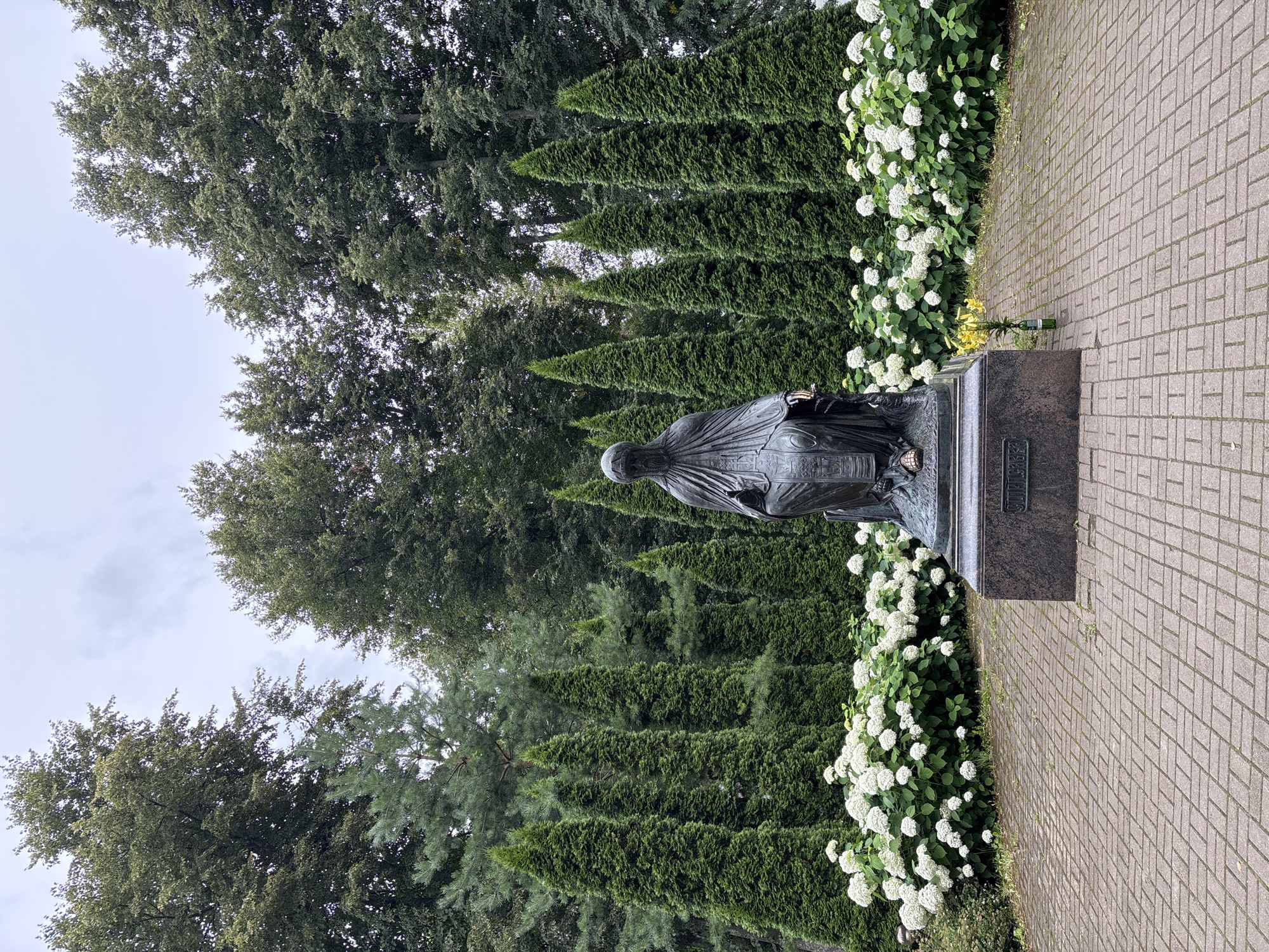 The image depicts a bronze statue of a seated cloaked figure on a granite pedestal. The statue is positioned centrally in a paved courtyard, surrounded by a semi-circular bed of white hydrangeas and tall, conical evergreen trees. In the background, a dense line of mature trees forms a backdrop under a cloudy sky. To the right of the statue's pedestal, a small bunch of yellow flowers in a green bottle is placed on the paving stones. The statue's pedestal has an inscription, which is partially visible. This scene is located at the Savvino-Storozhevsky Monastery in Zvenigorod, Russia.