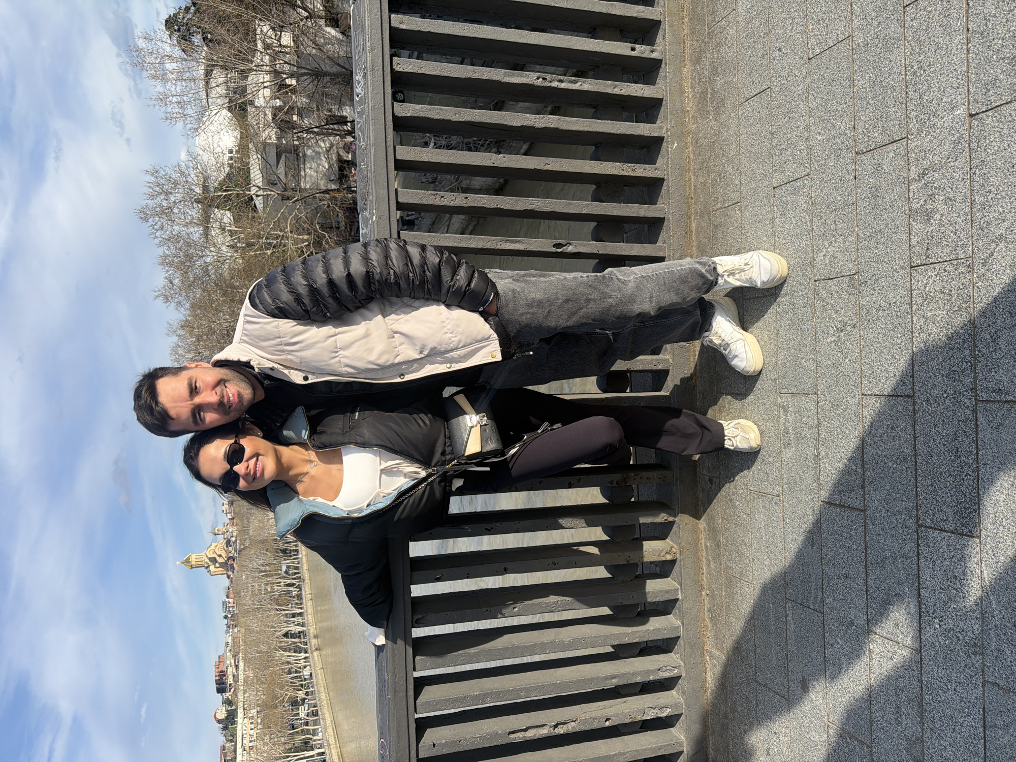 A man and a woman are photographed on a bridge, with the cityscape of Tbilisi, Georgia, visible in the background. The woman is wearing sunglasses, a black jacket, a white top, and black leggings, and she leans against the railing of the bridge. The man, standing behind her, wears a jacket and jeans. Both are smiling at the camera. The background shows buildings and a cathedral, with a bright, partly cloudy sky overhead. The bridge has a gray stone surface and a dark railing.