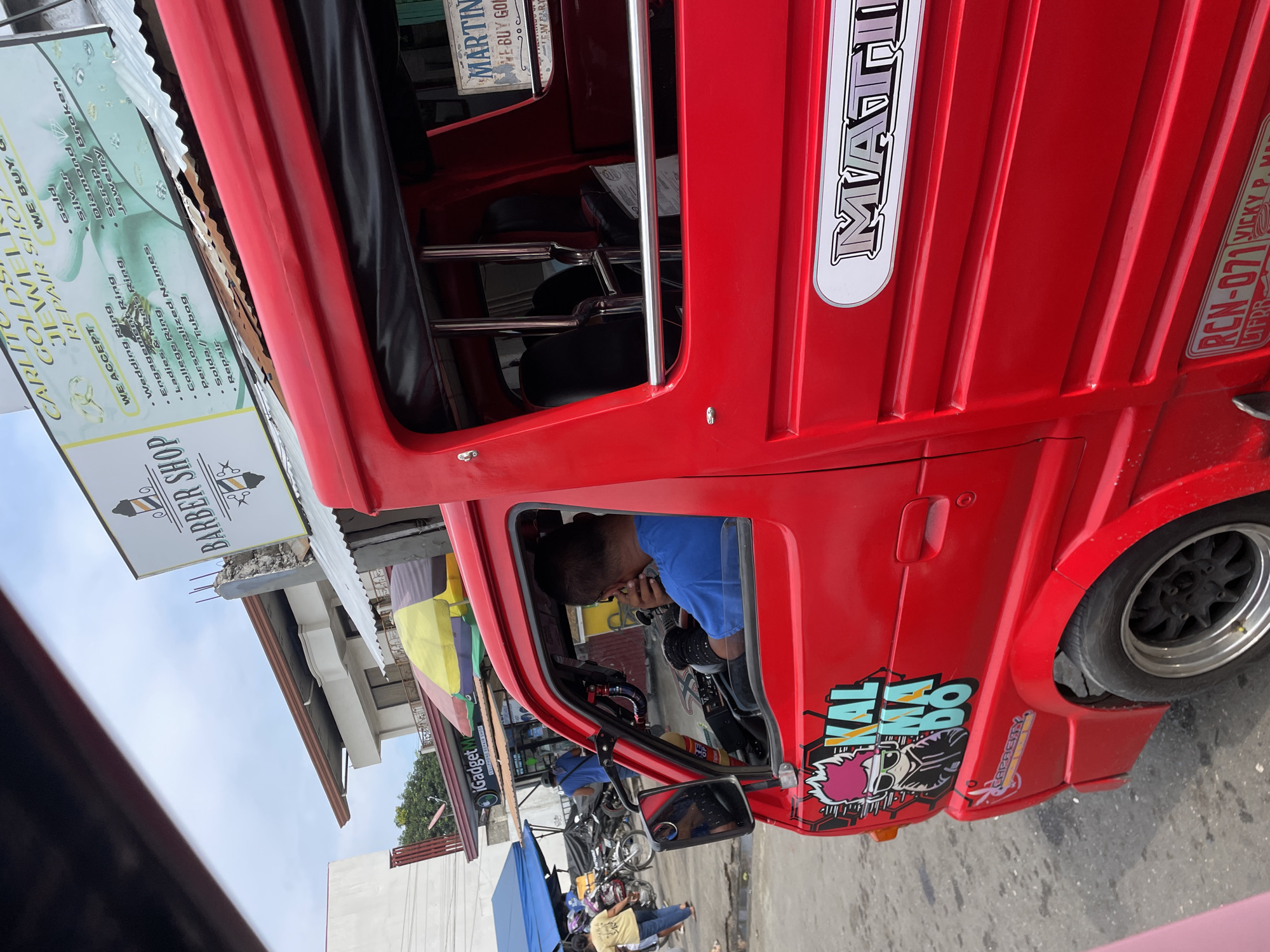 The image depicts a daytime urban street scene in Davao City, Philippines, captured from a slightly elevated perspective. The primary subject is a bright red, three-wheeled public utility vehicle, likely a motorized tricycle, occupying the lower and central right portion of the frame.
The red vehicle features a stylized "KAL MA DO" graphic with a pink-haired, sunglasses-wearing cartoon figure on its driver's side panel. A metallic "MATIN" logo is affixed to the rear passenger side. The lower rear indicates "RCN-071" and "VICKY P. MAR..." as part of its registration, with "LTFRB" visible. The vehicle has dark-colored rims with silver edges. Inside the driver's compartment, an adult male, wearing a blue short-sleeved shirt, is seated at the steering wheel, with his right hand raised towards his face. The passenger compartment, visible through an open window, shows black seats and chrome grab bars.
In the mid-ground, to the left of the red vehicle, an asphalt or dirt road surface is visible. Several people are present, including an individual in a yellow shirt and blue shorts walking, and another in a blue shirt further back. A cluster of motorcycles is parked near a blue tent. A commercial establishment named "iGadgetM" (likely "iGadget Mobile" or "iGadget Mart") with a "CELLPHONE REPAIR SHOP" sign is visible, flanked by yellow and green market umbrellas.
The background features commercial buildings. Prominently displayed are two large signs: a white "BARBER SHOP" sign with barber pole motifs, and a "CARLITO GOLD & JEWELRY SHOP" sign listing services such as "WE ACCEPT Wedding Ring, Engagement Ring, Ladies & Men's Ring, College Ring, Personalized Names, Solda/Tubog, Repair" and "WE BUY Gold, Silver, Diamond / Broken Jewelry." A smaller sign for "MARTIN" also indicates "WE BUY GOLD..." The sky above is partly cloudy.