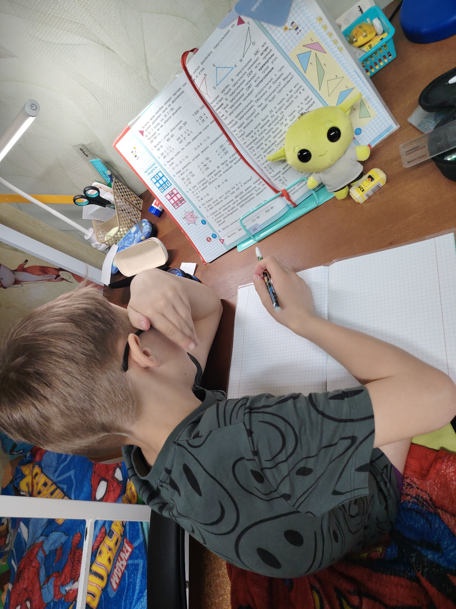 A boy with short, light brown hair is seated at a desk, working on his math homework. He is wearing a dark gray t-shirt with a smiley-face pattern. His left hand is covering his face, while his right hand holds a pen, poised to write in a graph-paper notebook. On the desk, next to the notebook, is an open textbook with mathematical problems written in Russian, along with a stuffed Grogu toy and various school supplies. The boy is sitting on a chair with a red and blue Spider-Man themed cover. A desk lamp illuminates the workspace. The setting appears to be a room in a private residence, likely Chaikovsky, Russia, given the Russian text in the textbook.