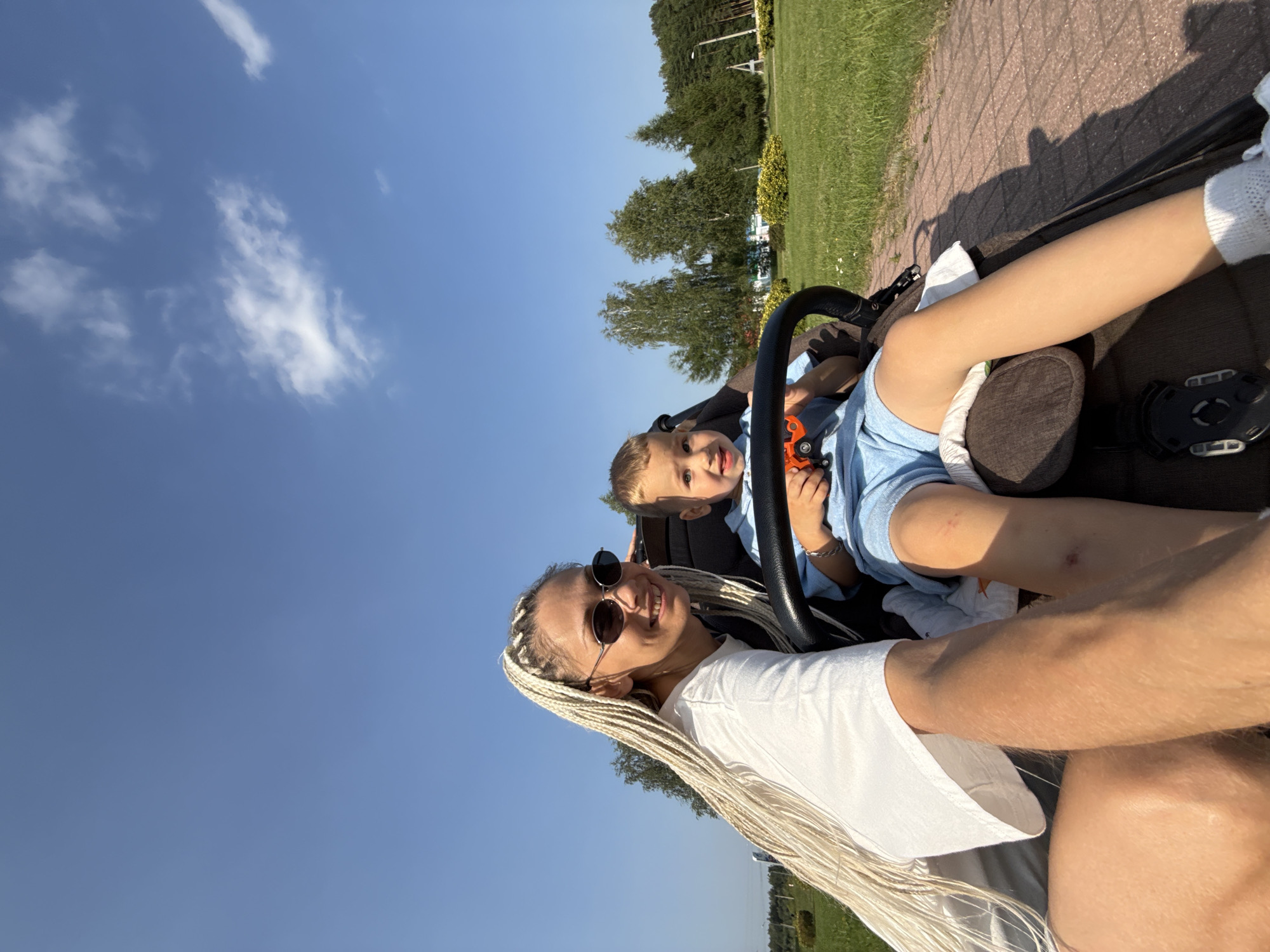 In Ignatovo, Russia, on a sunny day, a woman with long blonde braids and sunglasses smiles at the camera while sitting next to a young boy in a stroller. The boy, wearing a blue outfit, holds an orange toy. They are on a paved path, with a grassy area and trees in the background. The sky is clear blue with a few scattered clouds.