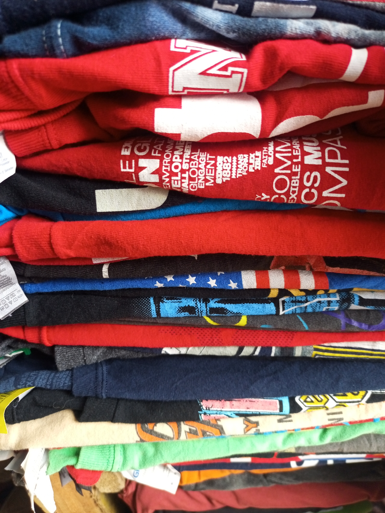The image shows a stack of folded clothing items. Predominantly red in color, the clothing has printed details, including lettering and graphic designs. Other colors present include blues, grays, blacks, yellows, greens, and oranges. One garment exhibits a U.S. flag design. The clothing appears to be casually folded and stacked on top of one another. The printing on the clothes includes words like "ENVIRONMENT," "GLOBAL," "ENGAGEMENT," "SINCE 1882," "FAITH," "FLEXIBLE LEARNING," "COMPASS," and "MUSIC," along with an "N" symbol. The garments have tags and fabric labels visible. The surrounding environment is out of focus, showing other stacked items and possibly cardboard.