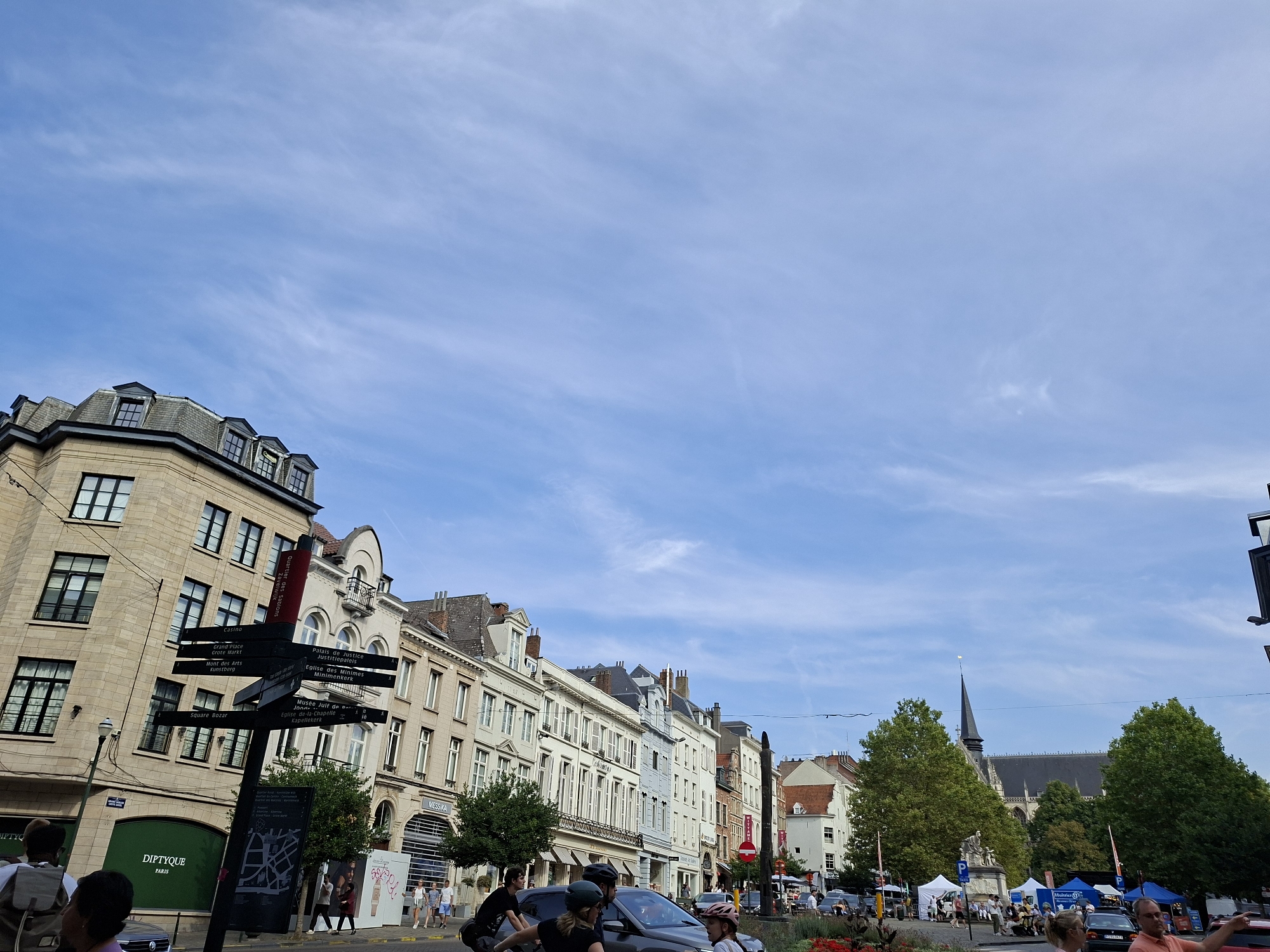 The image captures a street scene in Brussels, Belgium, likely in the vicinity of the Grand Place, given the signage. In the foreground, a black directional signpost points to various landmarks, including "Casino," "Grand Place," "Mont des Arts," and "Square Bazar." Several people are visible, some walking and others cycling, with a few cars also present on the street. The architecture features traditional European buildings with multiple stories and ornate facades. Trees line the street, and a church spire can be seen in the background. The sky is a clear blue with wispy white clouds, indicating fair weather. The overall impression is a bustling urban environment with a mix of historic and modern elements.
