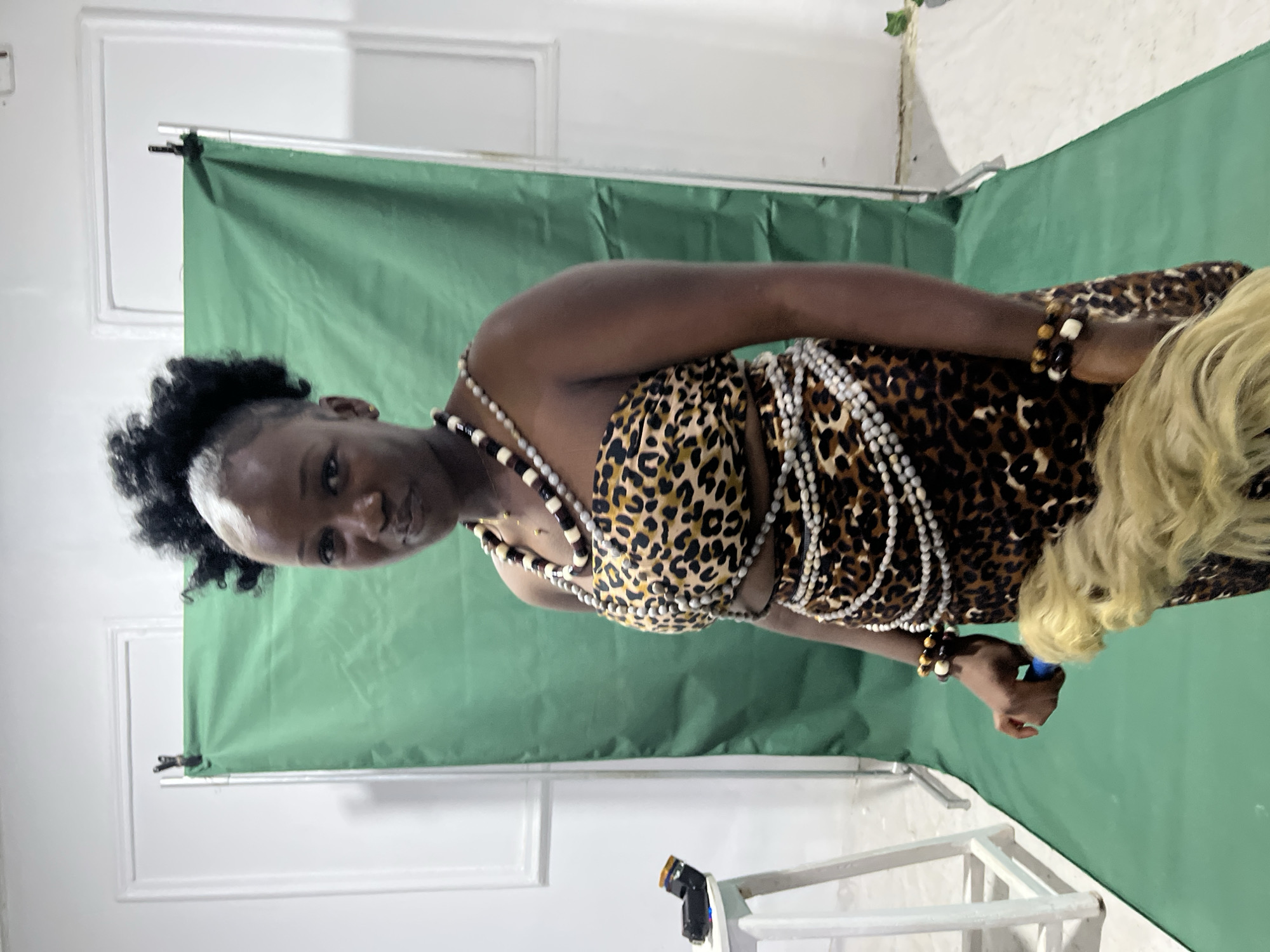 A dark-skinned woman stands in front of a green screen in an indoor setting. She is dressed in a leopard print outfit and adorned with multiple strands of white and dark brown beads. The background includes white walls and a white foldable chair. The setting suggests a photography studio or a makeshift studio setup. The location in the city of Jos, Nigeria is not discernible from the image alone.