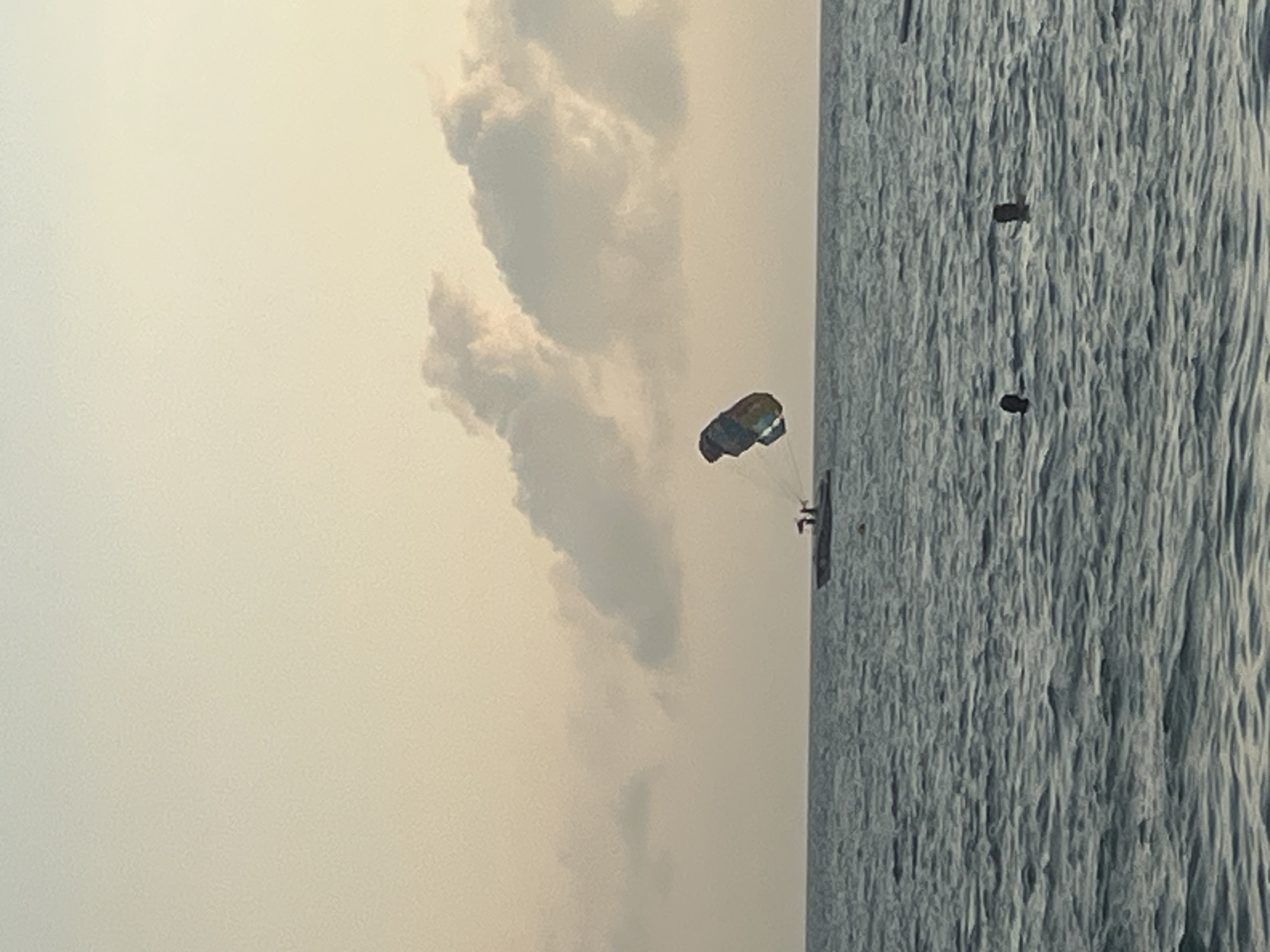 The image shows a parasailing activity on the sea. A boat is pulling a parachute with two people parasailing in the air above the water. The sky is overcast with soft, diffused light, suggesting it is either sunrise or sunset. The sea is calm with gentle ripples. In the foreground, two people are swimming in the water, with their heads just above the surface. The scene is set against a pale, hazy sky. This scene could be taking place in the city of Sochi, Russia, a popular Black Sea resort known for its beaches and water sports.
