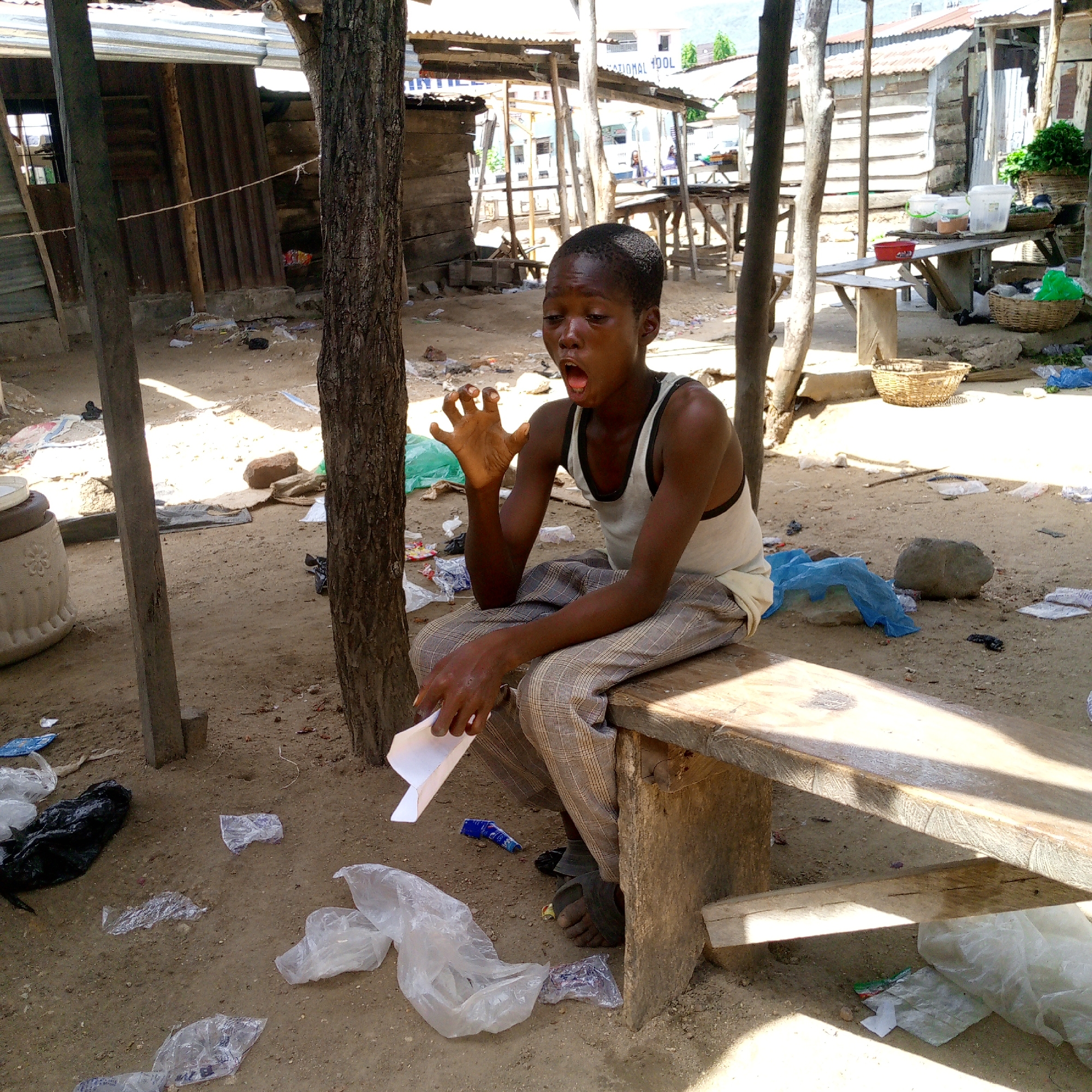 This image depicts a young boy sitting on a rough wooden bench in an outdoor, unpaved area in Adankolo, Nigeria, under bright daylight. The boy, who appears to be a pre-teen or young teenager, has dark skin and short hair. He is wearing a white tank top with dark trim and plaid trousers, along with dark sandals. His mouth is wide open in a dramatic expression, possibly a yawn, shout, or gasp, while his left hand is raised with fingers splayed. He holds a folded piece of white paper or cardboard in his right hand, resting his arm on his knee.
The scene is characterized by a dusty dirt ground, heavily littered with numerous plastic bags, wrappers, and other debris. Surrounding the boy are various makeshift structures, predominantly constructed from wooden planks and corrugated metal sheets, which serve as walls and roofs for stalls or dwellings. In the mid-ground, several rudimentary wooden tables or market stalls are visible, some holding wicker baskets, plastic containers, and fresh produce.
The strong, overhead sunlight creates distinct shadows, suggesting it is mid-day. In the far background, a larger, more structured white building can be seen, partially obscured by other elements. The text "NATIONAL POOL" is faintly visible on the side of this building. The overall setting portrays an informal, bustling, and somewhat unkempt market or community area, reflecting a basic living or commercial environment. The boy's expressive face is the emotional focal point of the photograph.