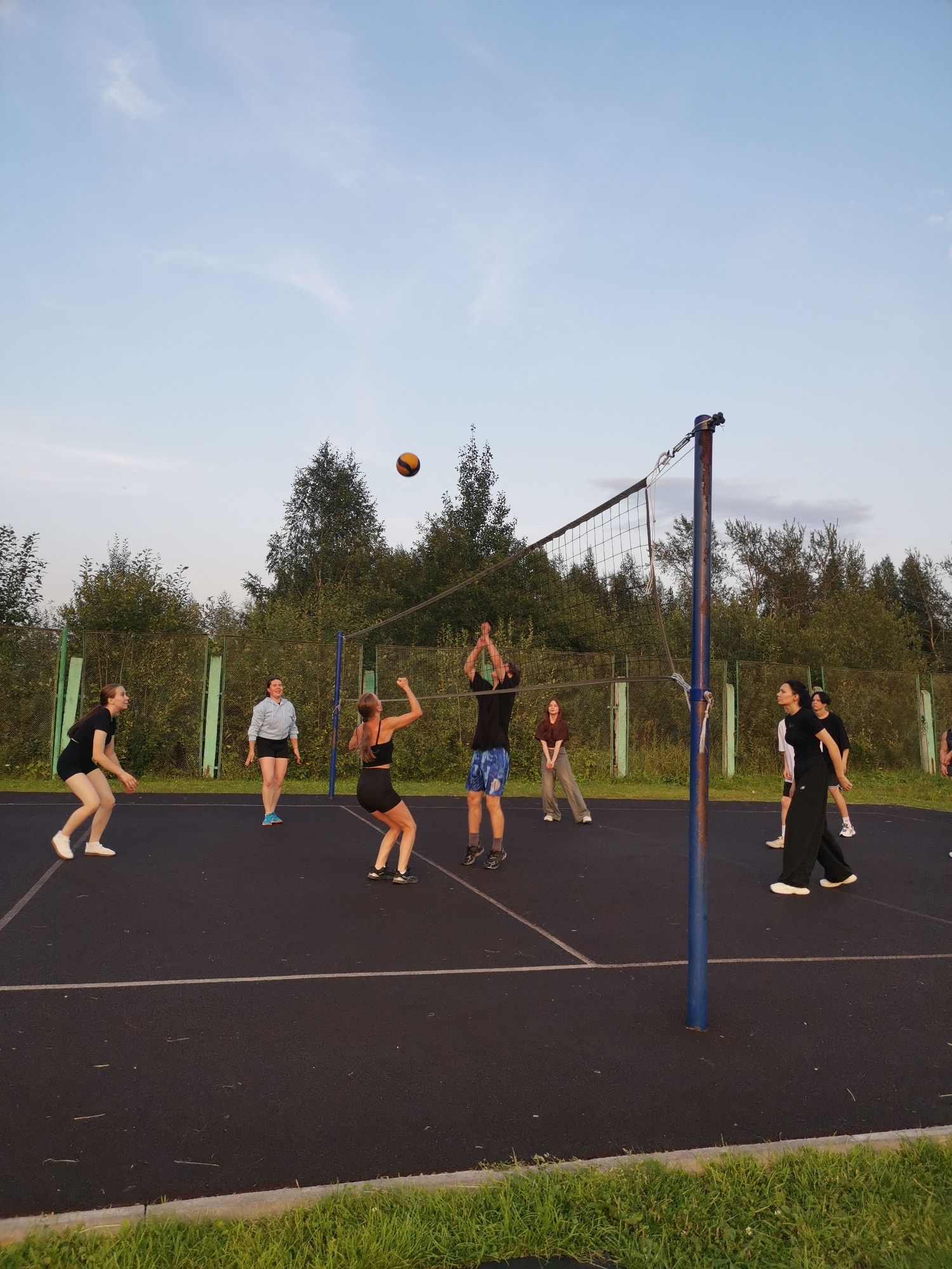 This image depicts a group of eight individuals playing volleyball on an outdoor court. The court is black asphalt with white lines marking boundaries. A blue pole supports a volleyball net, which is strung across the court. The background consists of a green fence, dense trees, and a pale blue sky with some wispy clouds.
Seven people are actively engaged in the game. One person in the center is jumping to hit the ball, while another person on the left is in a defensive stance, ready to receive. Another person is in the process of serving or hitting the ball, with arms raised. Three other individuals are positioned on the court, likely awaiting their turn or observing the play. One person is standing further back, partially obscured by the net.
The individuals are dressed in casual athletic wear, including shorts, t-shirts, and sports bras. Their actions suggest a dynamic game in progress. The lighting indicates it is likely late afternoon or early evening, with the sun casting soft light. The environment appears to be a recreational area, possibly in a park or school. The city of Kizel, Russia is not visually identifiable within the image.