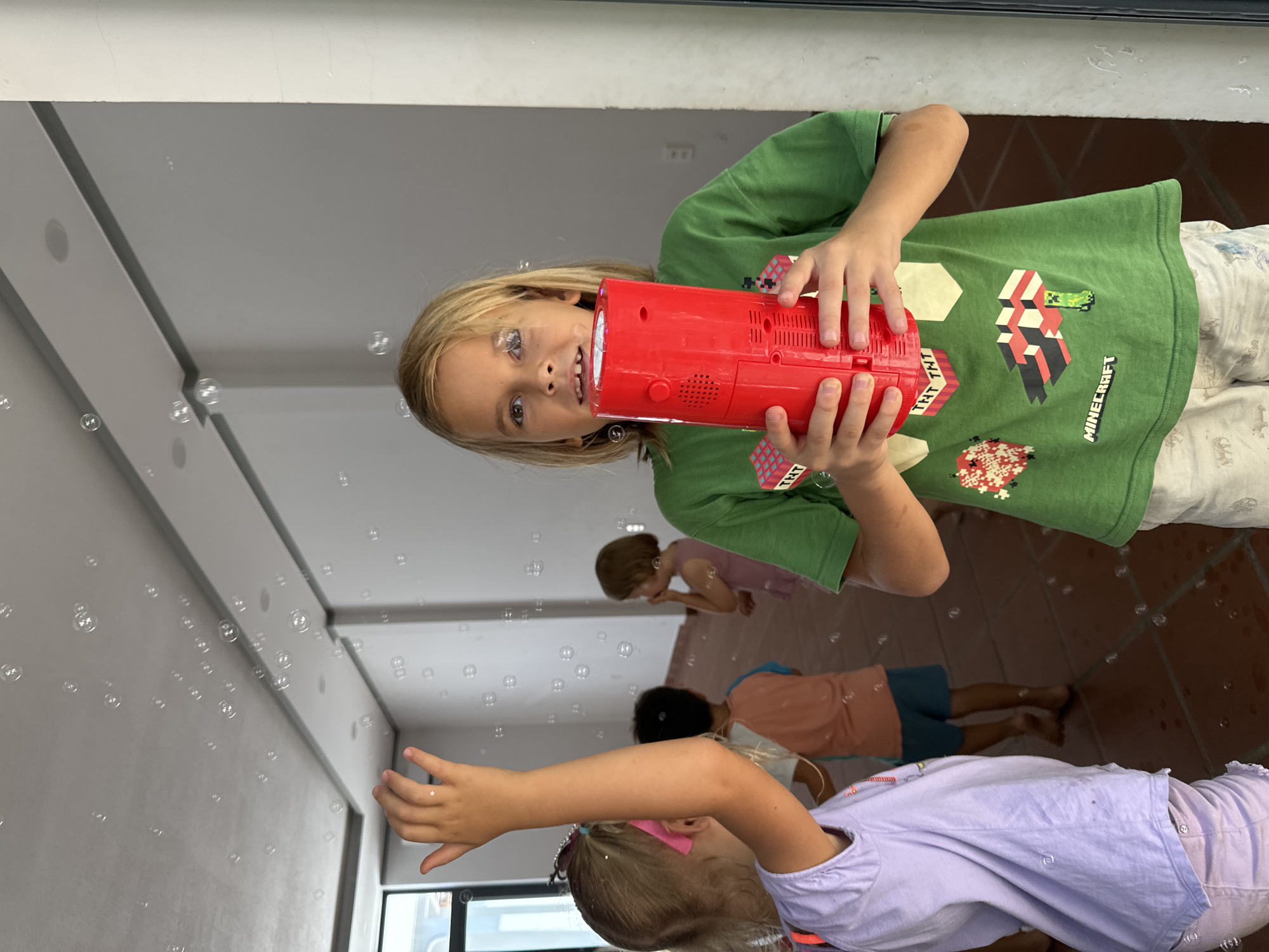 The image captures a lively scene indoors or in a covered outdoor area, likely in Mũi Né, Vietnam, centered around children playing with bubbles. The primary subject is a child with long blonde hair and light-colored eyes, positioned prominently in the foreground. This child is smiling brightly and looking towards the viewer while holding a vibrant red bubble machine with both hands. The child is wearing a green t-shirt that clearly displays the text "MINECRAFT" and "TNT" graphics.
Numerous transparent bubbles fill the air around the children, indicating an active play session. In the mid-ground and background, several other children are visible, engaging with the bubbles. One child, wearing a pink top, appears to be bending down, possibly to pop bubbles or collect them from the reddish-brown tiled floor. Another child, further back, wears a peach or orange top and blue shorts, also in a crouched or kneeling position. Partially visible on the right side of the frame is the arm and head of a child in a lavender top and a pink headband, reaching out towards the floating bubbles.
The setting features light-colored walls or a ceiling, possibly with reflective surfaces or architectural elements, suggesting a bright and spacious environment. The overall atmosphere is joyful and playful, conveyed by the children's engagement and the abundance of bubbles. The lighting indicates it is daytime.