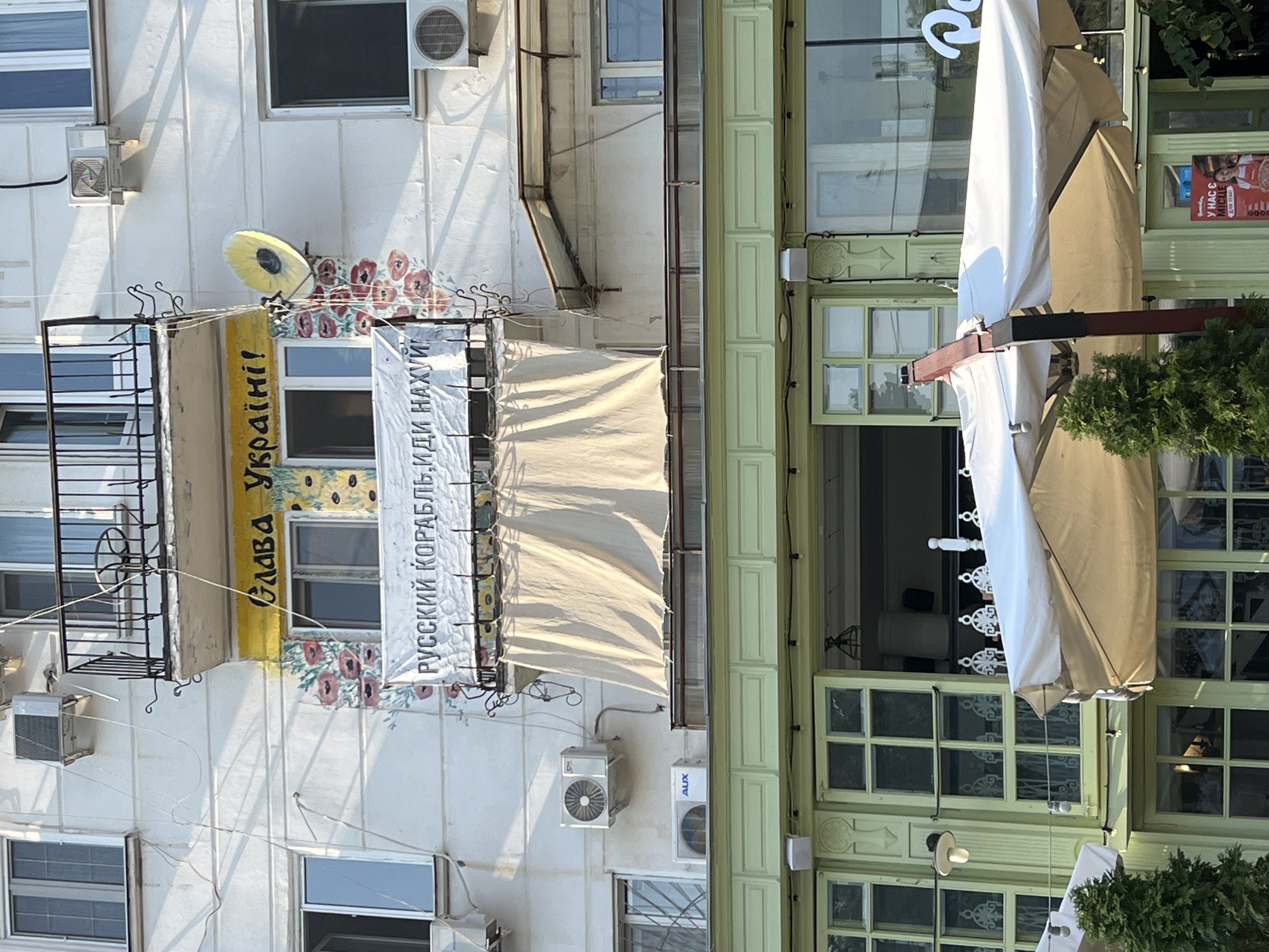 The image depicts a facade of a building in Odesa, Ukraine, featuring an anti-Russian slogan painted on the wall and a banner. The slogan reads "Слава Україні!" (Glory to Ukraine!) in yellow letters on a yellow background, accompanied by floral imagery of poppies and sunflowers. Below this, a white banner with black Cyrillic text states "РУССКИЙ КОРАБЛЬ, ИДИ НАХУЙ" (Russian warship, go fuck yourself). A satellite dish is mounted on the wall next to the slogan.
The building has multiple windows and air conditioning units visible. A green-paneled structure, possibly a cafe or restaurant with large windows and outdoor seating areas covered by umbrellas, is situated below the building. The overall scene appears to be a street view in Odesa, with the graffiti and banner conveying a political message in response to the ongoing conflict.