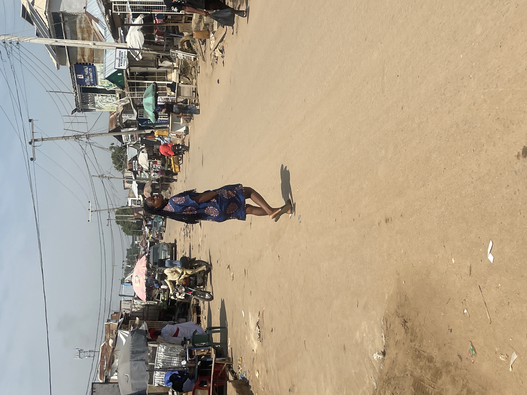 In Abuja, Nigeria, a street scene unfolds under a hazy sky. A woman in a blue and patterned dress walks away from the camera down a dirt road lined with bustling market stalls and shops. Various people are visible in the background, some walking, others on motorcycles. A man in a yellow outfit rides a red motorcycle, and another person in a red shirt walks near a stall. Buildings with corrugated metal roofs and visible electrical wires dominate the sides of the road. The ground is a dusty, sandy terrain with scattered litter, including a plastic bag and paper fragments. The overall impression is of a vibrant, active marketplace on a bright, possibly warm day.