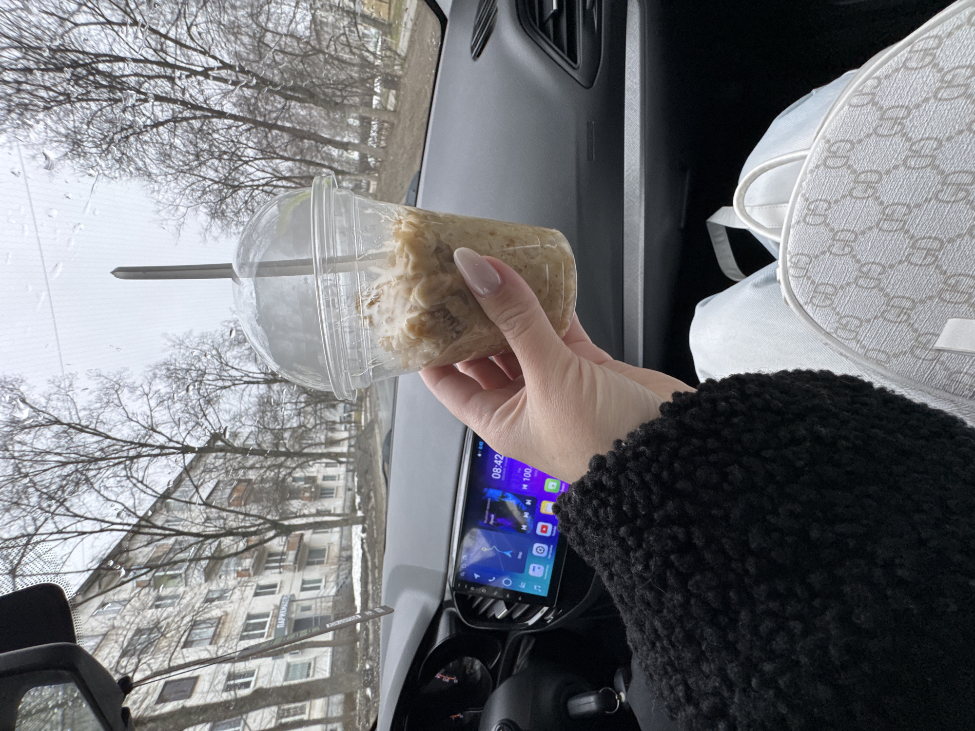 A hand holds a transparent plastic cup containing a beige, granular substance, possibly food, inside a car. The cup is covered with a lid and has a straw. The car's dashboard is visible with a central screen displaying navigation and other applications, showing a time of 08:42. The exterior view through the windshield shows a gray, overcast sky, bare trees, and a residential building. The building appears to be of Soviet-era construction, with multiple windows and a balcony. There is a sign in Russian script on the building that reads "ПАРКМАША." Considering the context of the sign, the location is likely in the city of Chaikovsky, Russia, as the street name appears on the sign. The presence of a luxury bag is also visible within the vehicle, positioned in the passenger seat. The person holding the cup is wearing a black sweater with a distinct texture. Raindrops are present on the windshield.
