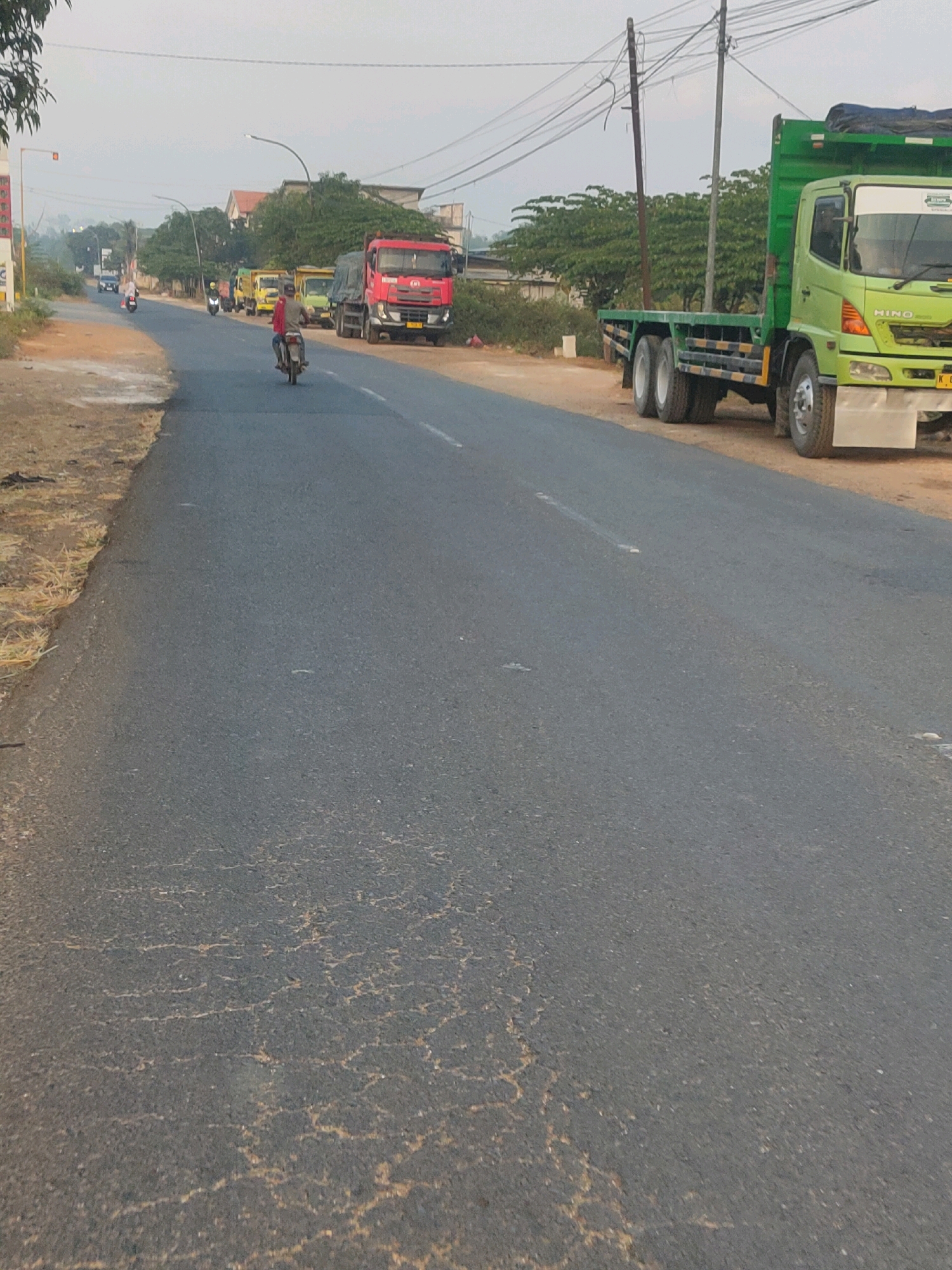 The image depicts a rural roadside scene in Sawo, Indonesia, characterized by a paved road with a cracked surface. A person is riding a motorcycle in the foreground, moving away from the viewer. Several trucks, including a bright red one and a green flatbed truck, are parked or moving along the road. The surrounding environment features vegetation, utility poles with visible wires, and buildings in the background, suggesting a moderately developed area. The sky is overcast.