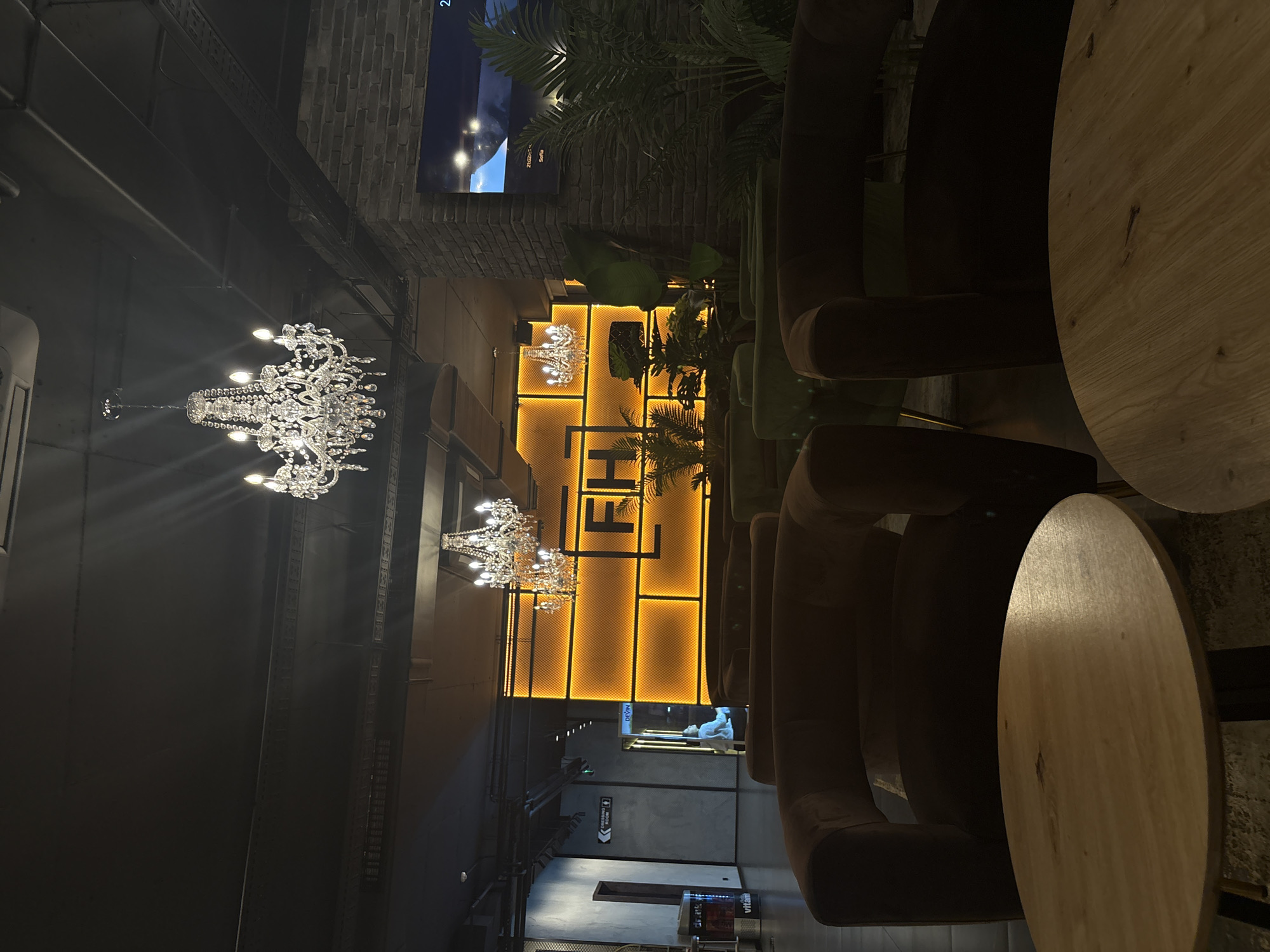 The image depicts the interior of a stylish establishment, likely a cafe, restaurant, or lounge, in Plovdiv, Bulgaria. The setting is dimly lit, suggesting evening or nighttime, and relies heavily on artificial lighting to create ambiance.
**Scene and Setting:** The space features a contemporary design, blending industrial elements with luxurious touches. Dark walls and ceilings are prominent, with exposed pipes or structural beams visible. A striking feature is a large, back-lit wall display with a warm, golden-yellow glow, forming a grid pattern and incorporating the large, stylized letters "FH". Lush green plants are strategically placed, adding a natural element.
**Objects:**
* **Lighting:** Two ornate, multi-tiered crystal chandeliers hang from the ceiling, providing bright, sparkling light. The "FH" wall feature is significantly illuminated. A television screen displaying "23:02" and text like "Sofa" is visible in the upper background.
* **Furniture:** Several plush, dark-colored (possibly brown or velvet) armchairs are arranged around round, light wooden tables, suggesting comfortable seating for patrons.
* **Decor:** Besides the chandeliers and plants, sections of a brick wall are visible, contributing to the mixed aesthetic. Exposed metal structures and dark paneling are also present.
**People:** One person is faintly visible in the deeper background, near the restrooms, appearing to be standing.
**Activity:** No specific activity is taking place; the image is an interior shot showcasing the venue's design and atmosphere.
**Notable Details:**
* **Time of Day:** Implied to be evening or night due to the reliance on artificial light and the "23:02" timestamp on the screen.
* **Color Palette:** Dominated by dark tones for walls and furniture, contrasted with the warm yellow glow of the "FH" feature, the bright sparkle of the chandeliers, and the natural green of the plants and light wood of the tables.
* **Location Cues:** While the image itself doesn't show Plovdiv, Bulgaria, the context provided specifies it.
* **Text:** Visible text includes "FH" on the main backlit wall, "23:02" and "Sofa" on the TV screen, "GENTS & LADIES ROOM" on a directional sign, and "DKYN" and "vitamin" on another sign further in the background.